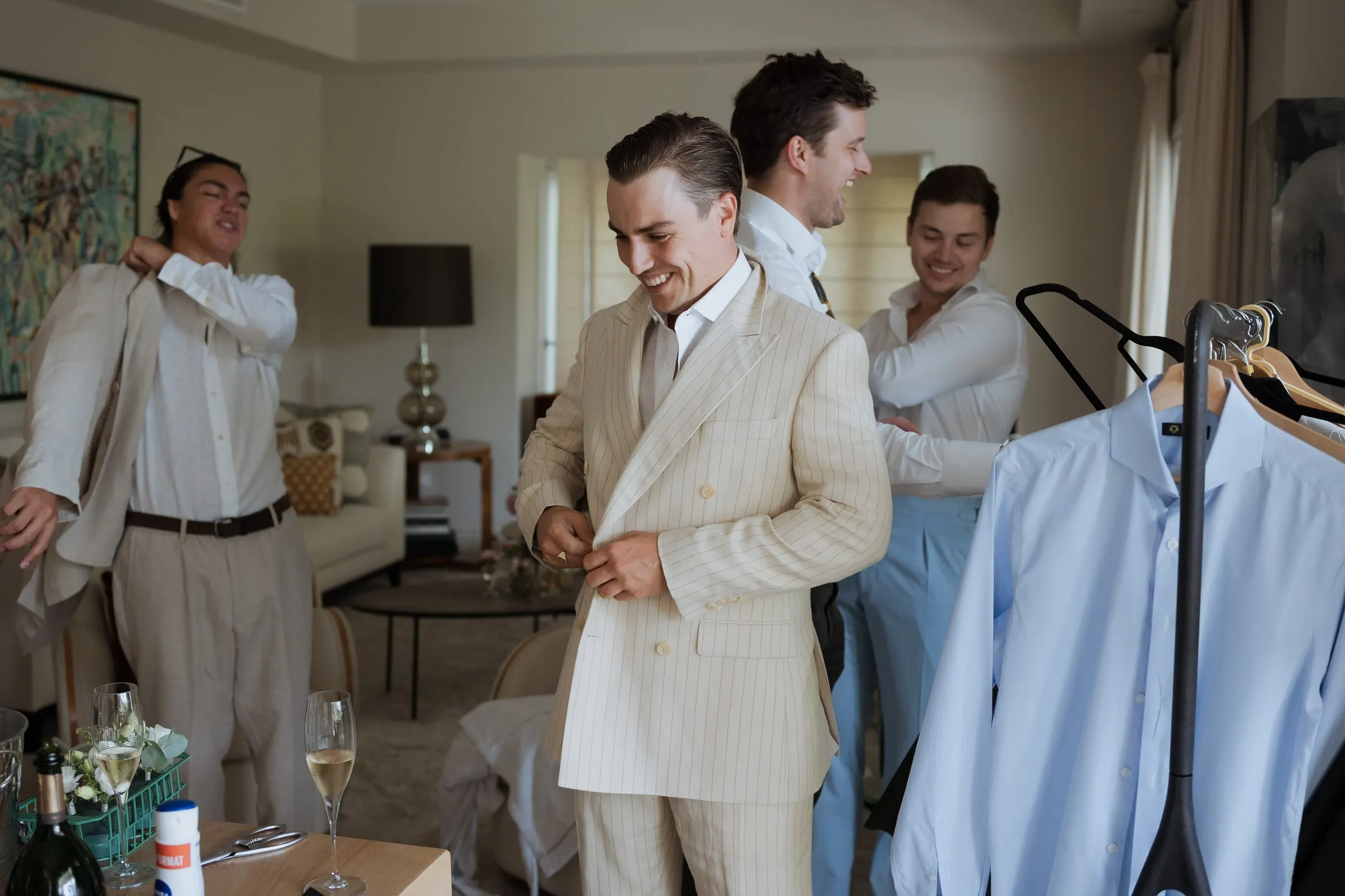 Groom laughing as he buttons his cream suit among friends getting dressed, Barbillón, Marbella
