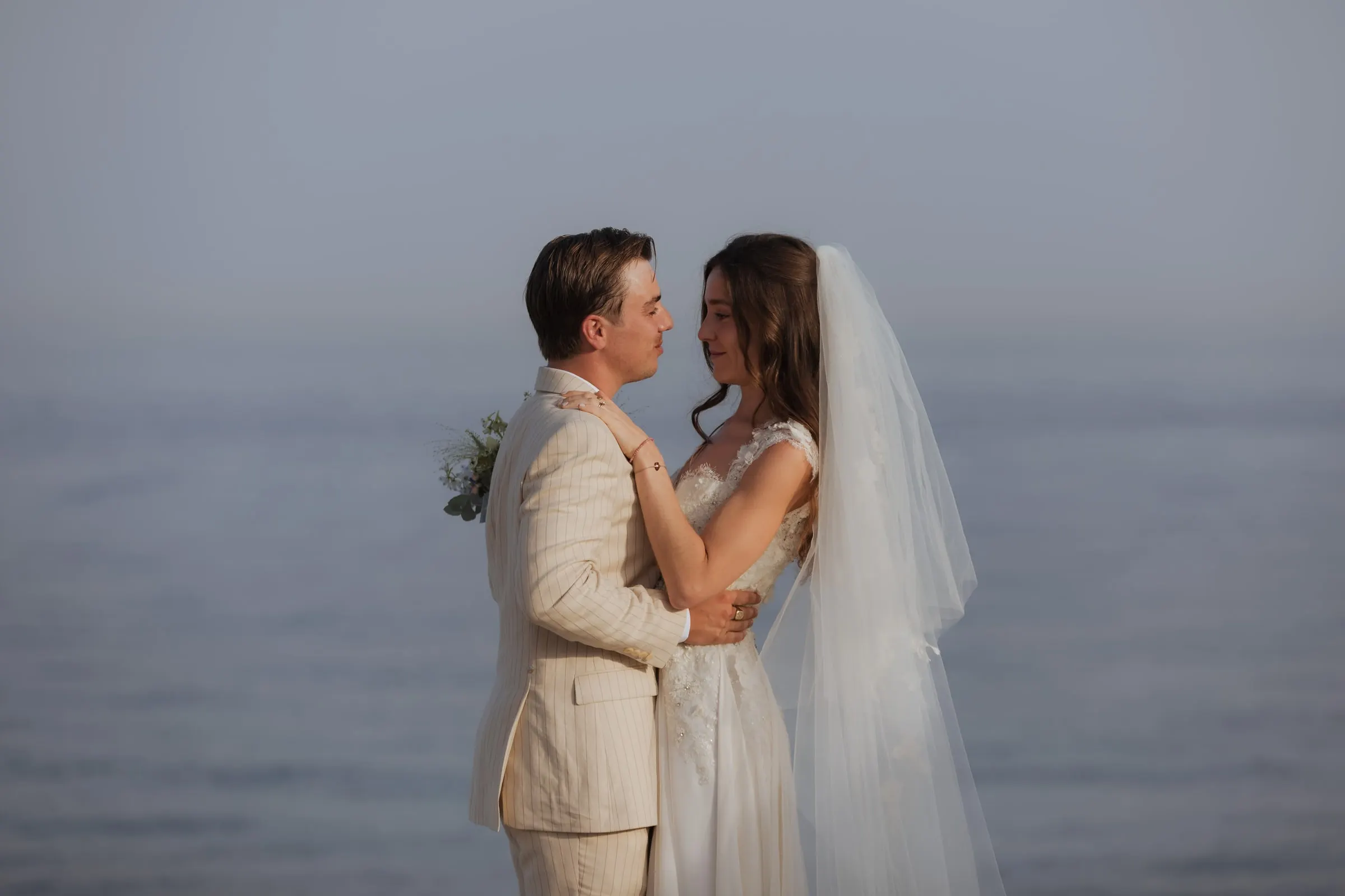 Bride and groom embracing in front of the sea, her veil in the breeze, Barbillón, Marbella