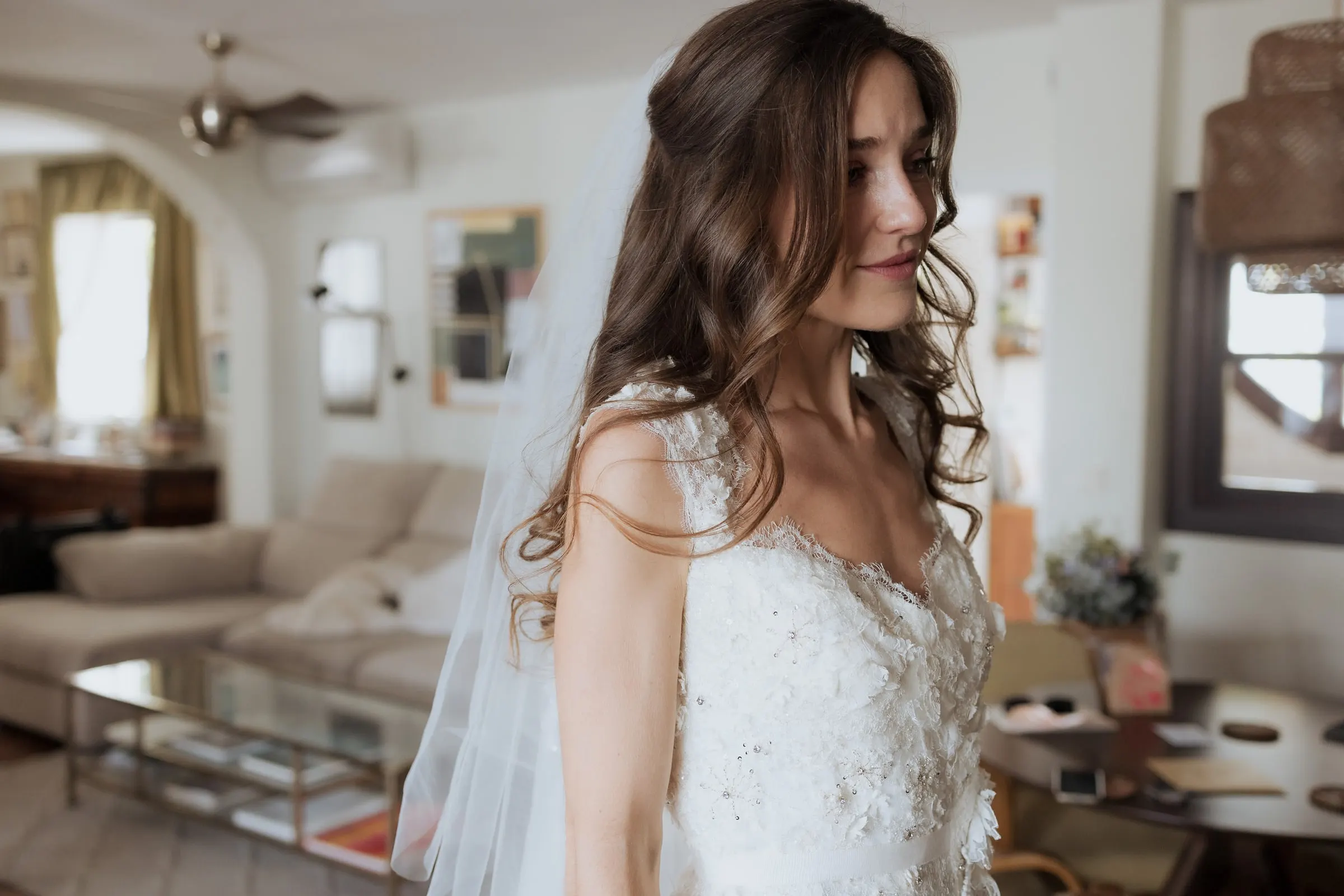 Bride with long wavy hair and a veil looking down in a bright living room before her wedding at Barbillón, Marbella