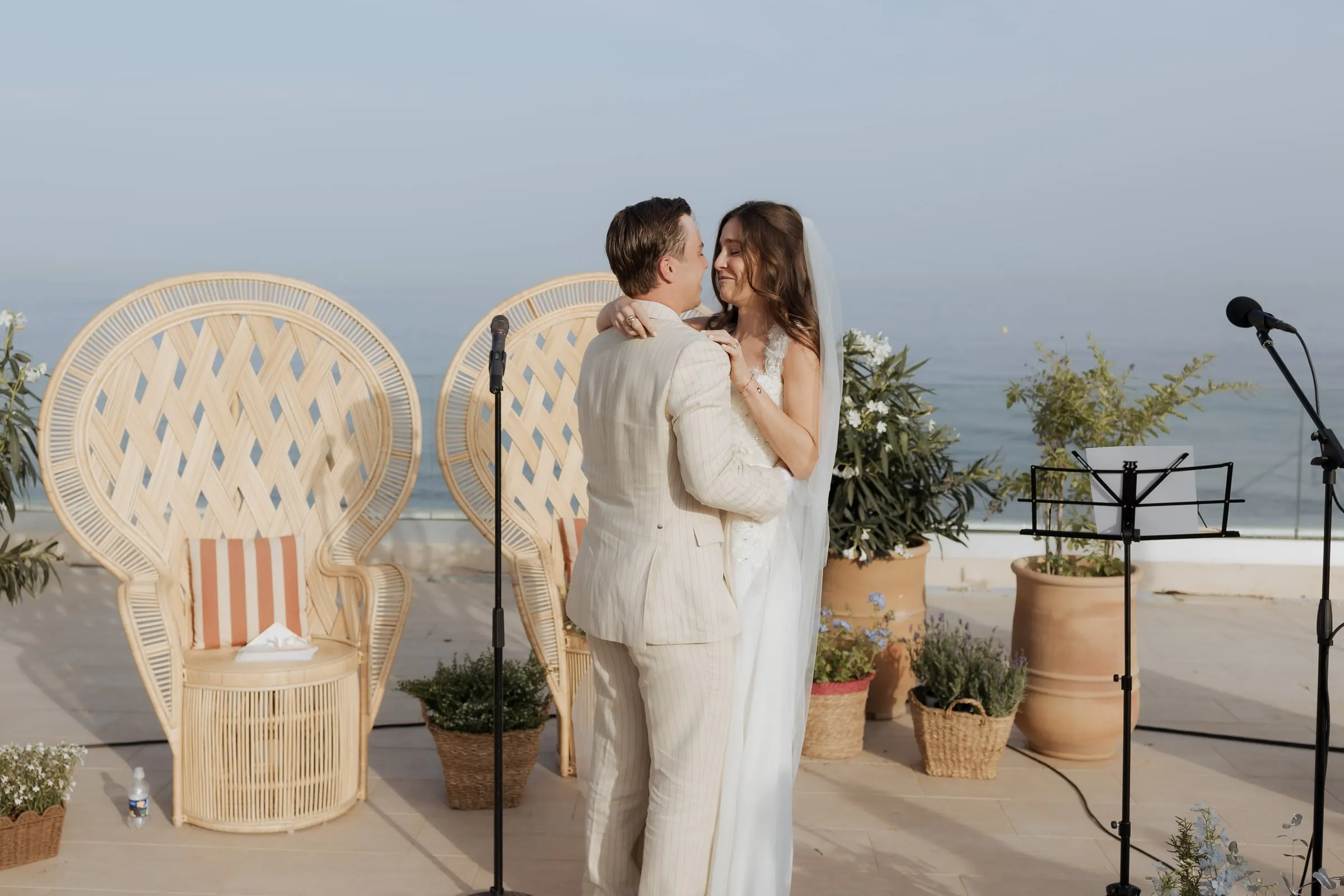 Bride and groom embracing between wicker peacock chairs and microphone stands, with the sea behind, at Barbillón, Marbella