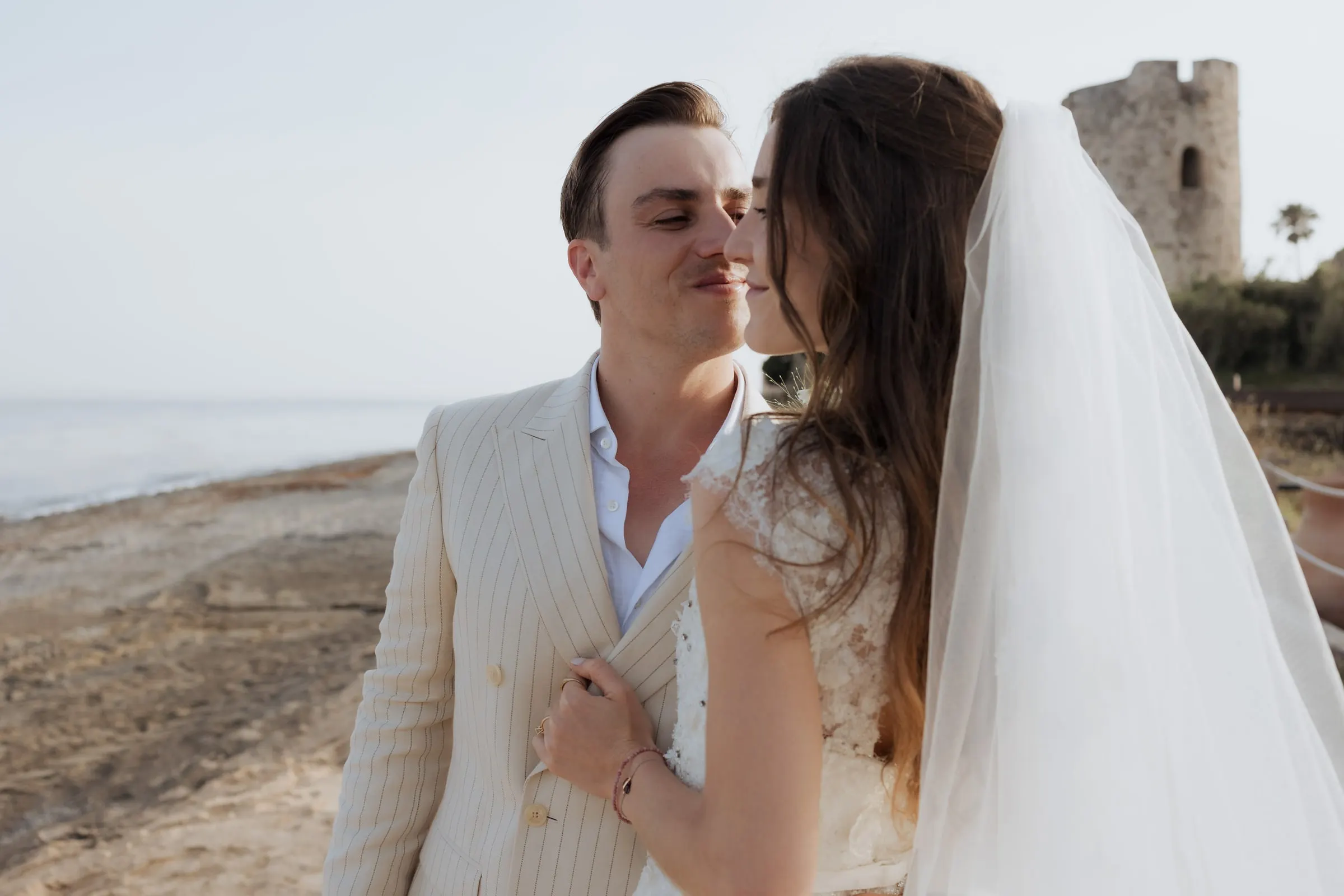 Close-up of the bride and groom about to kiss, with the sea and a stone watchtower behind, at Barbillón, Marbella