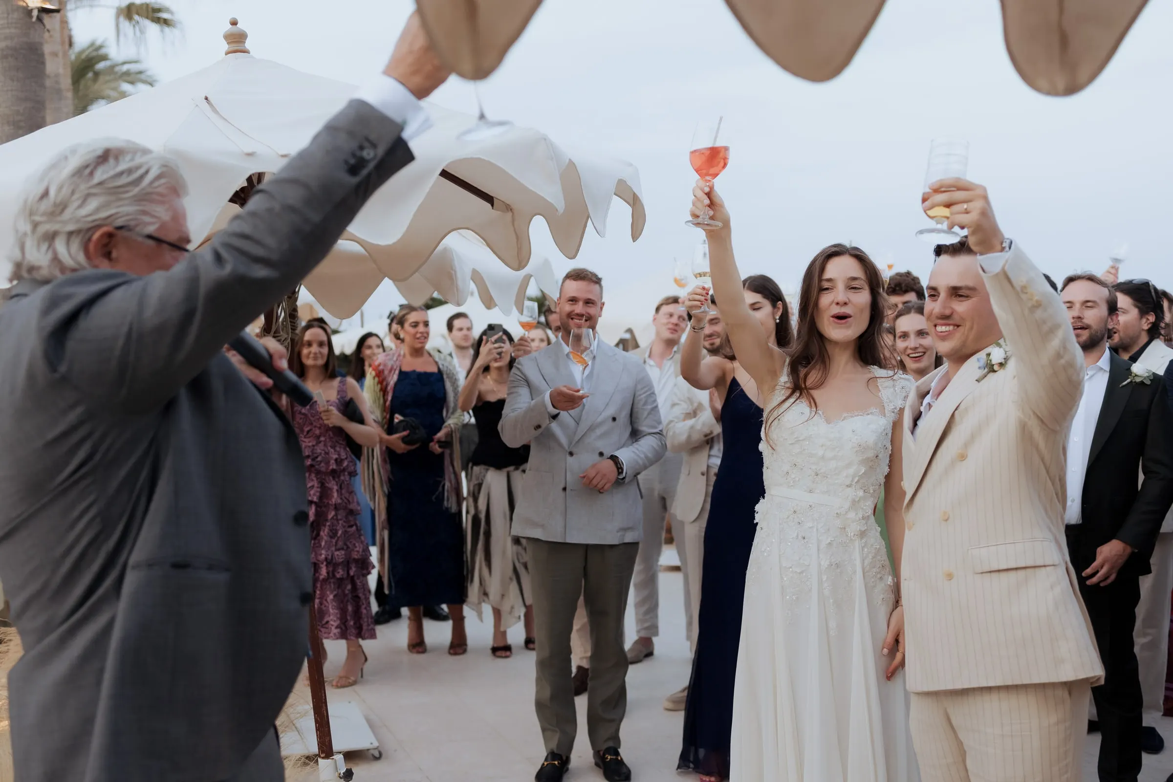 Bride and groom raising their glasses with guests under white umbrellas at Barbillón, Marbella