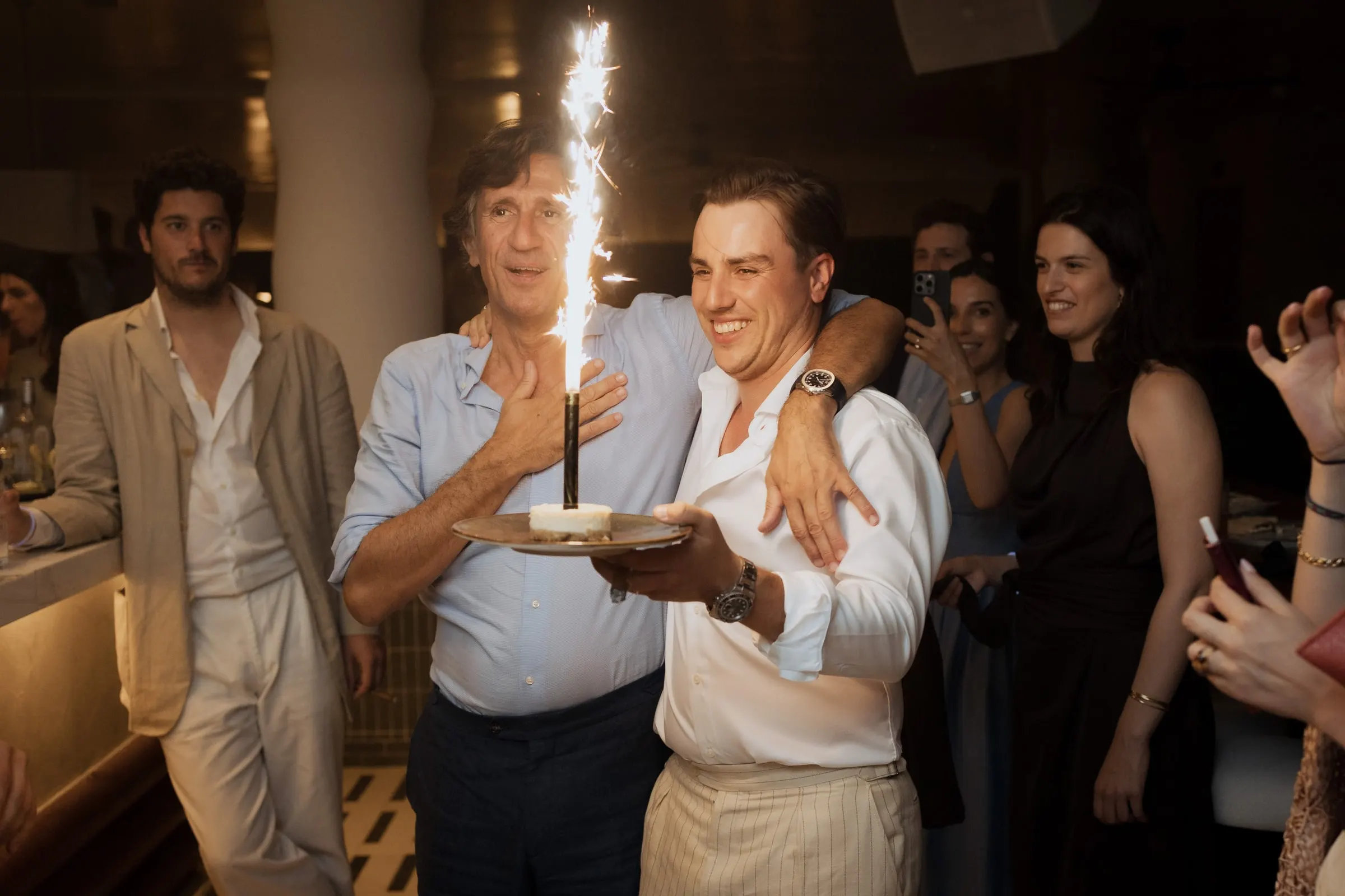 Groom carrying a plate with a sparkler candle as a guest puts his arm around him at the party at Barbillón, Marbella