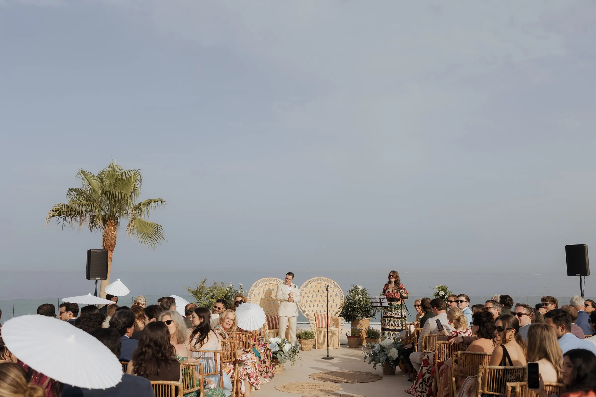 Wide view of the seafront ceremony with seated guests and white parasols, Barbillón, Marbella