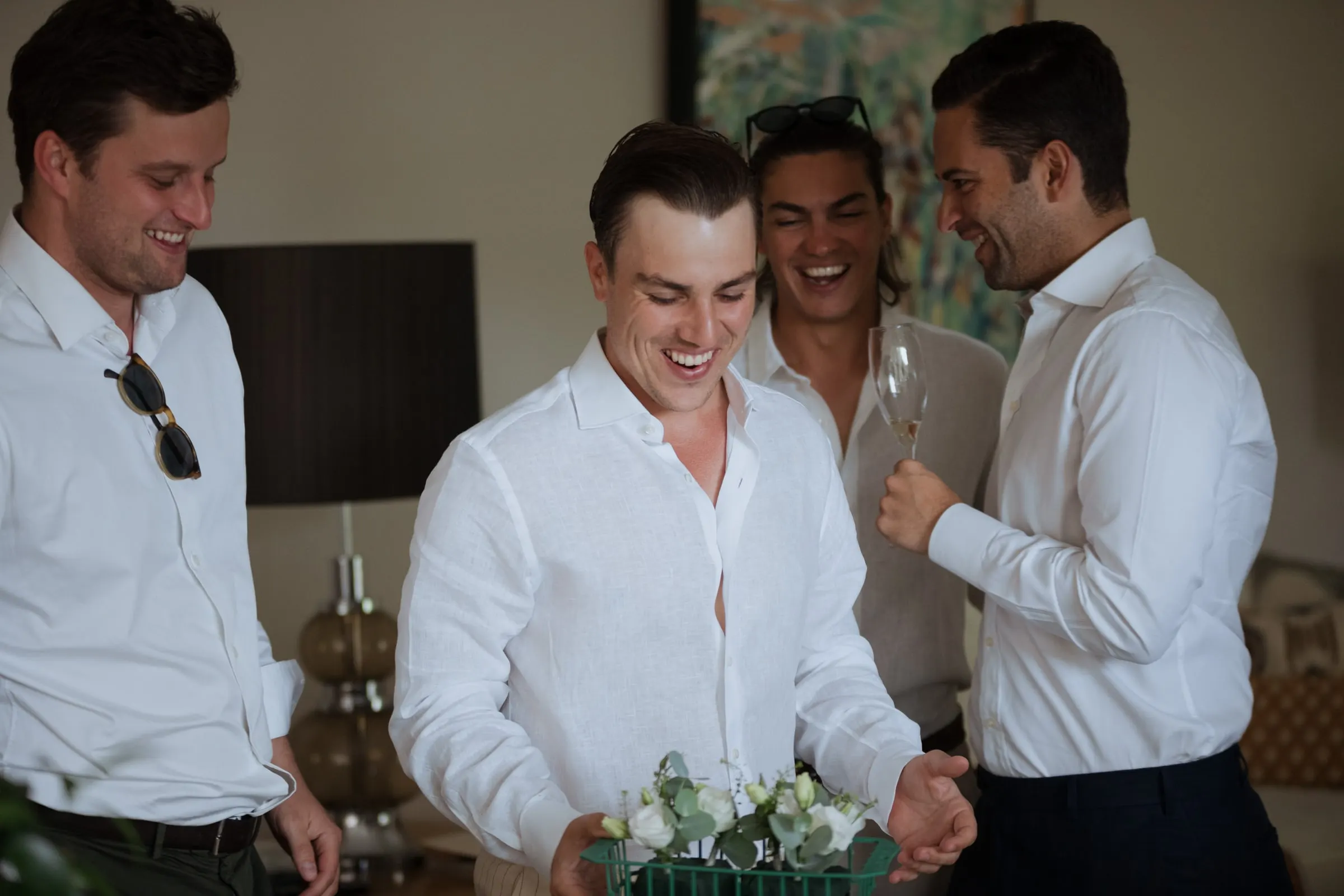 Groom laughing with a box of buttonholes surrounded by friends with drinks, Barbillón, Marbella