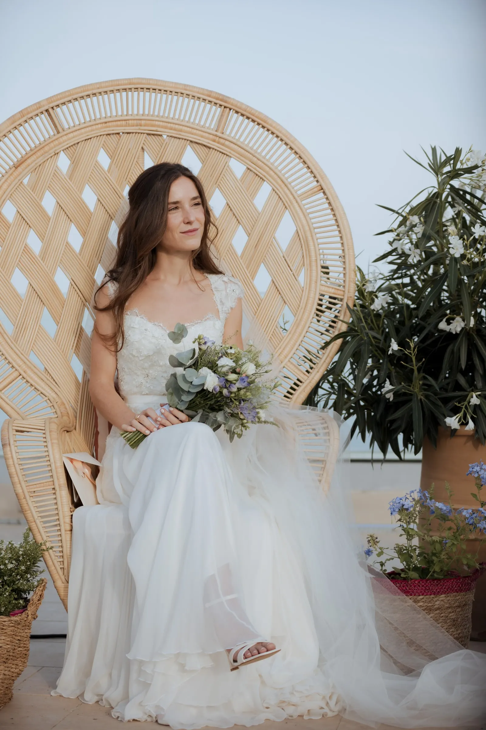 Bride seated in a wicker peacock chair during the ceremony by the sea, Barbillón, Marbella