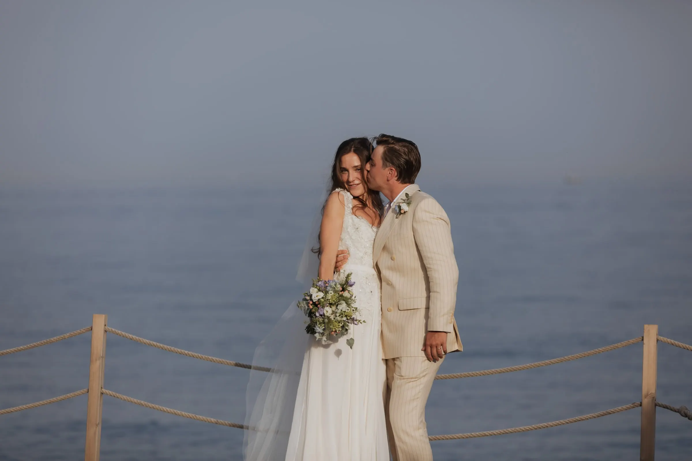 Groom kissing the bride's cheek by a rope railing above the sea, Barbillón, Marbella