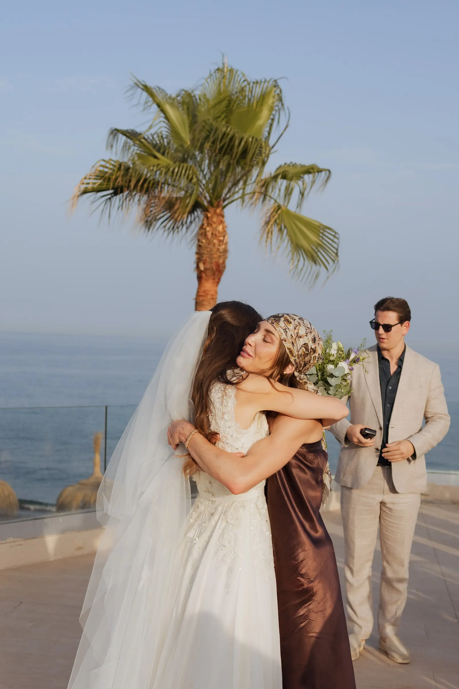 Bride hugging a guest in a brown dress on the terrace under a palm, Barbillón, Marbella