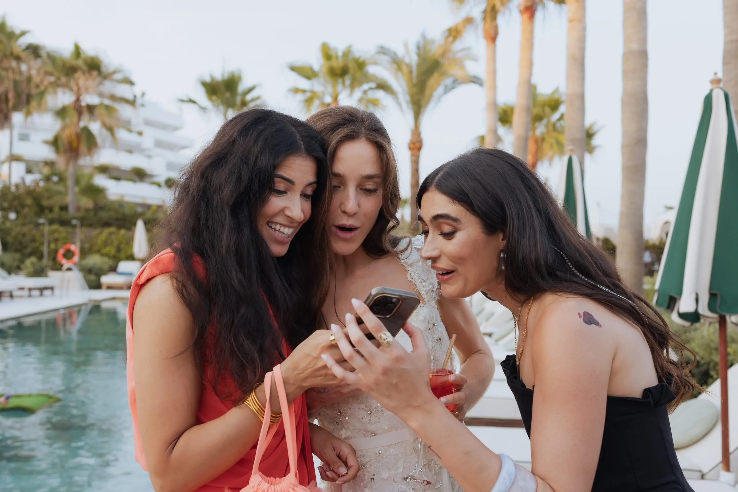 La novia y dos amigas ríen mirando un móvil junto a la piscina, Barbillón, Marbella