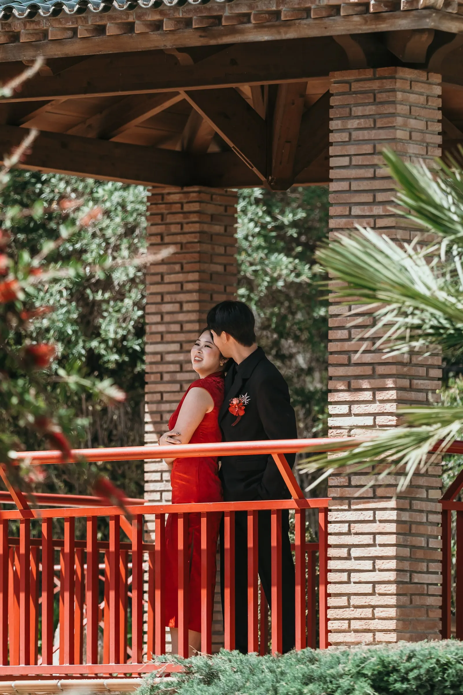 Couple leaning together by a brick pillar and the red railing of the pagoda