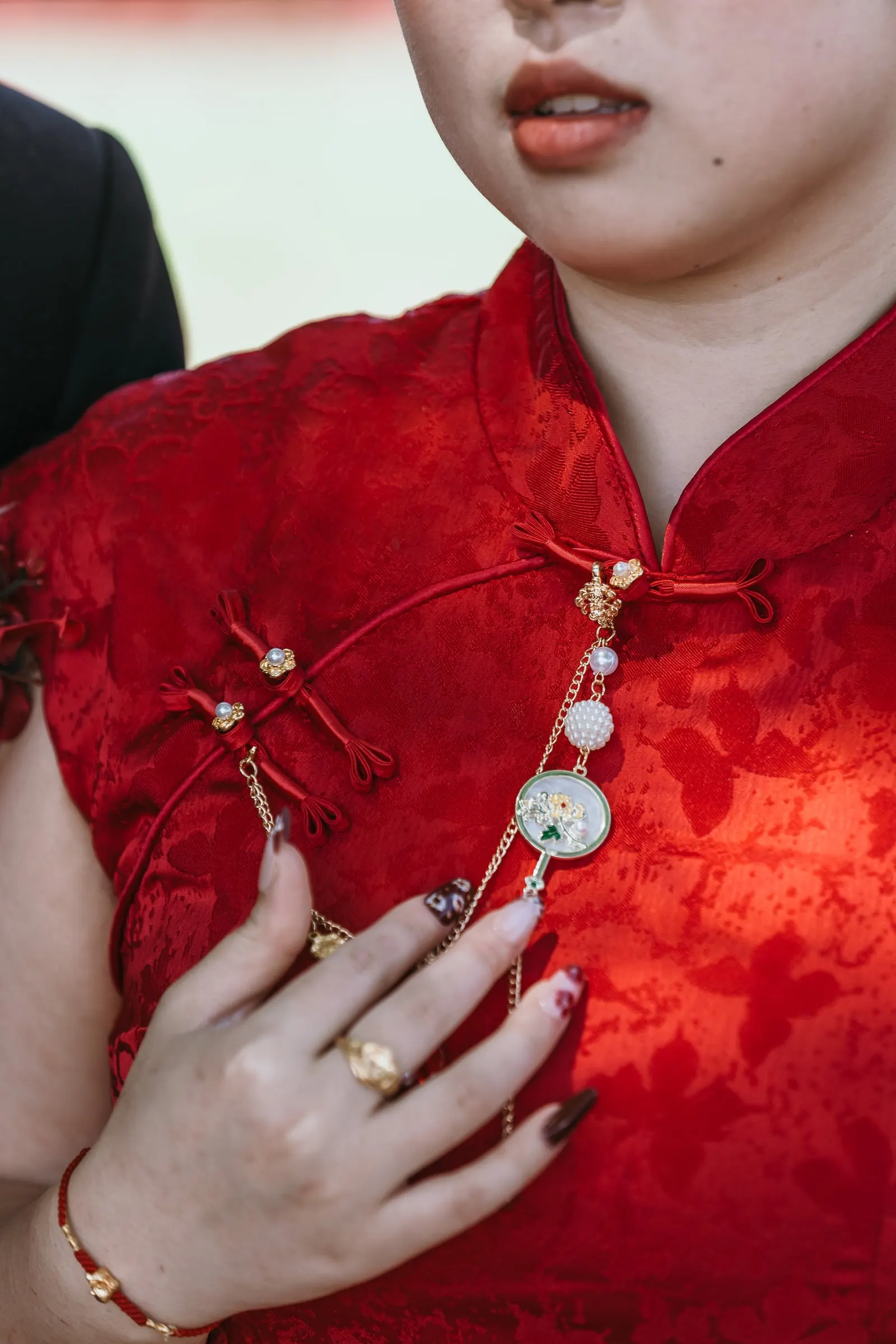 Detail of her hand on the red qipao with frog buttons, a pendant chain, a gold ring and bracelet