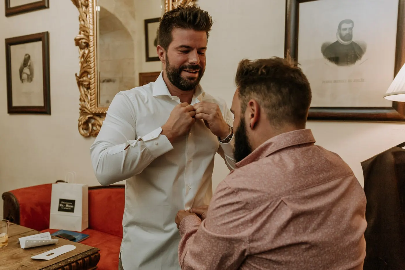 Groom buttoning his white shirt among old portraits and a red sofa, Castillo del Buen Amor