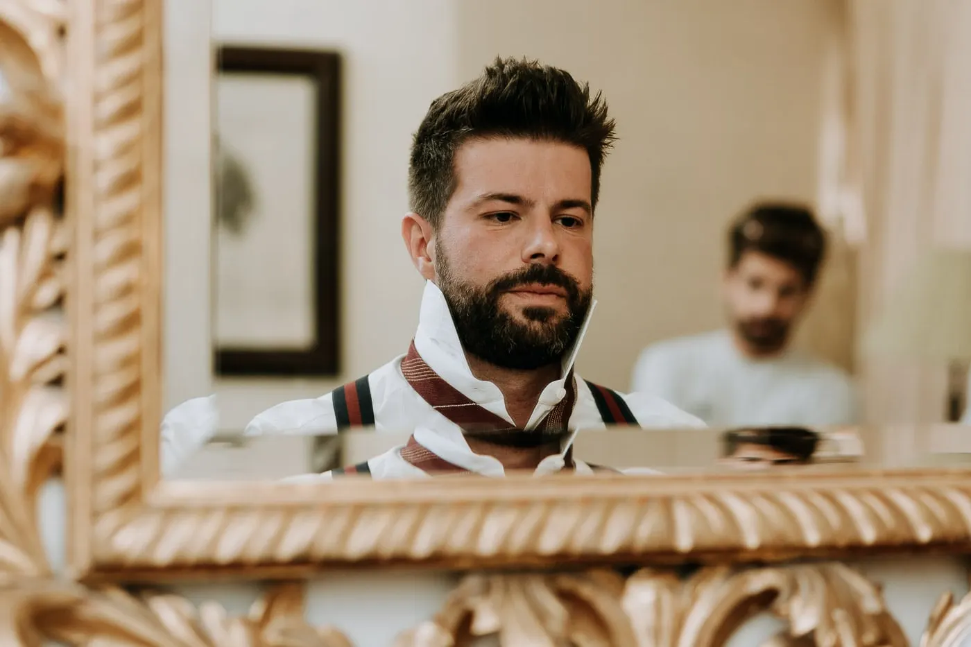 Groom tying his burgundy tie, reflected in a gilded mirror, Castillo del Buen Amor