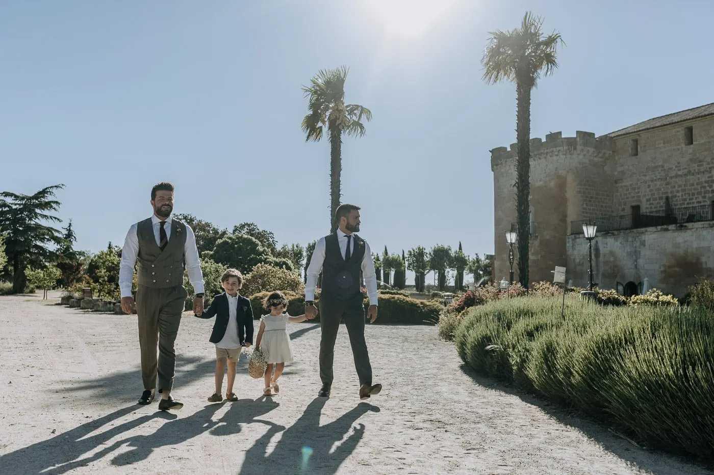 Grooms and children walking down the palm-lined drive towards the ceremony, Castillo del Buen Amor