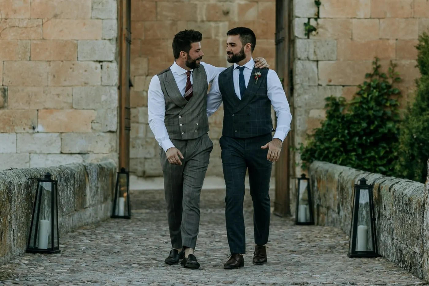 The grooms walking arm in arm along the lantern-lined stone path, Castillo del Buen Amor