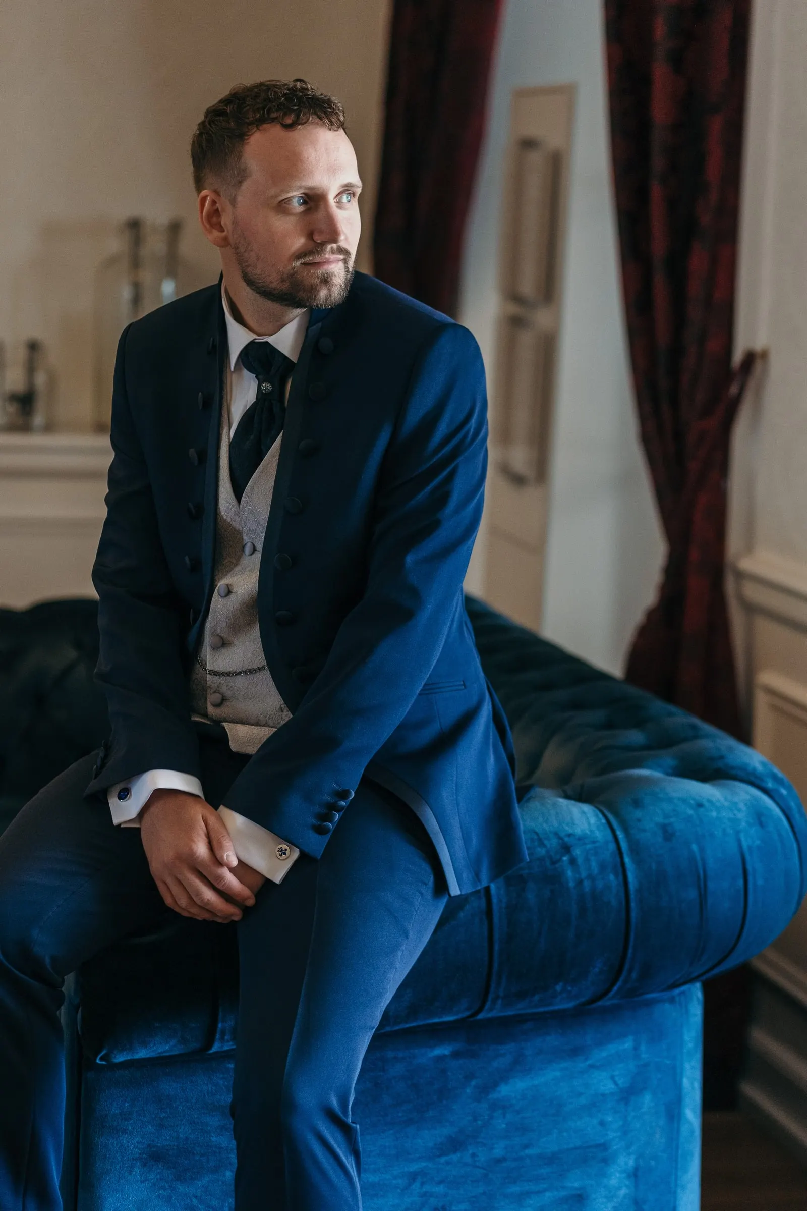 Vincent, con traje azul marino, sentado en un sofá de terciopelo azul de su habitación antes de la boda