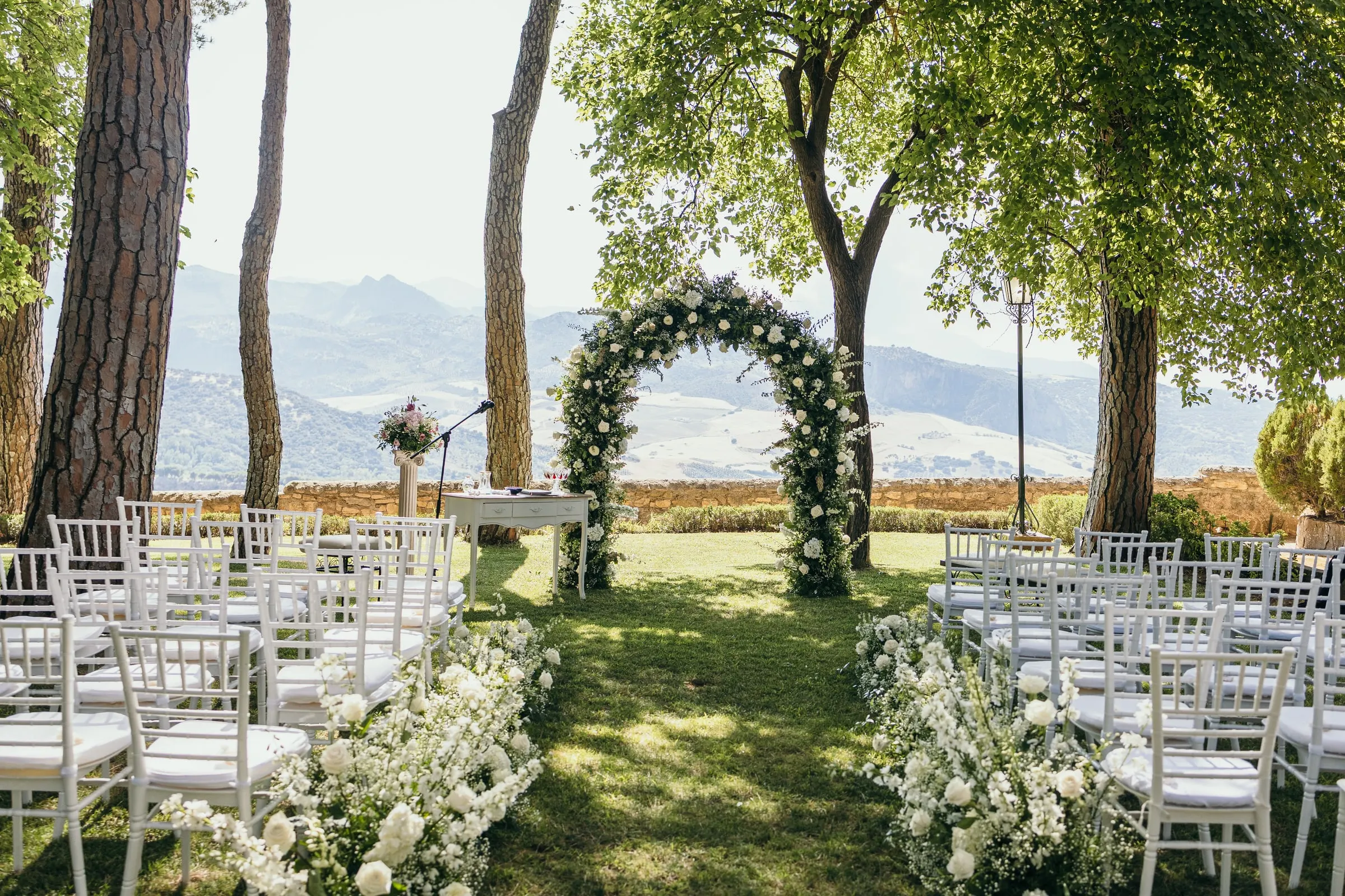 White chairs and a white flower arch under the pines of the Reina Victoria gardens, with the Serranía de Ronda behind