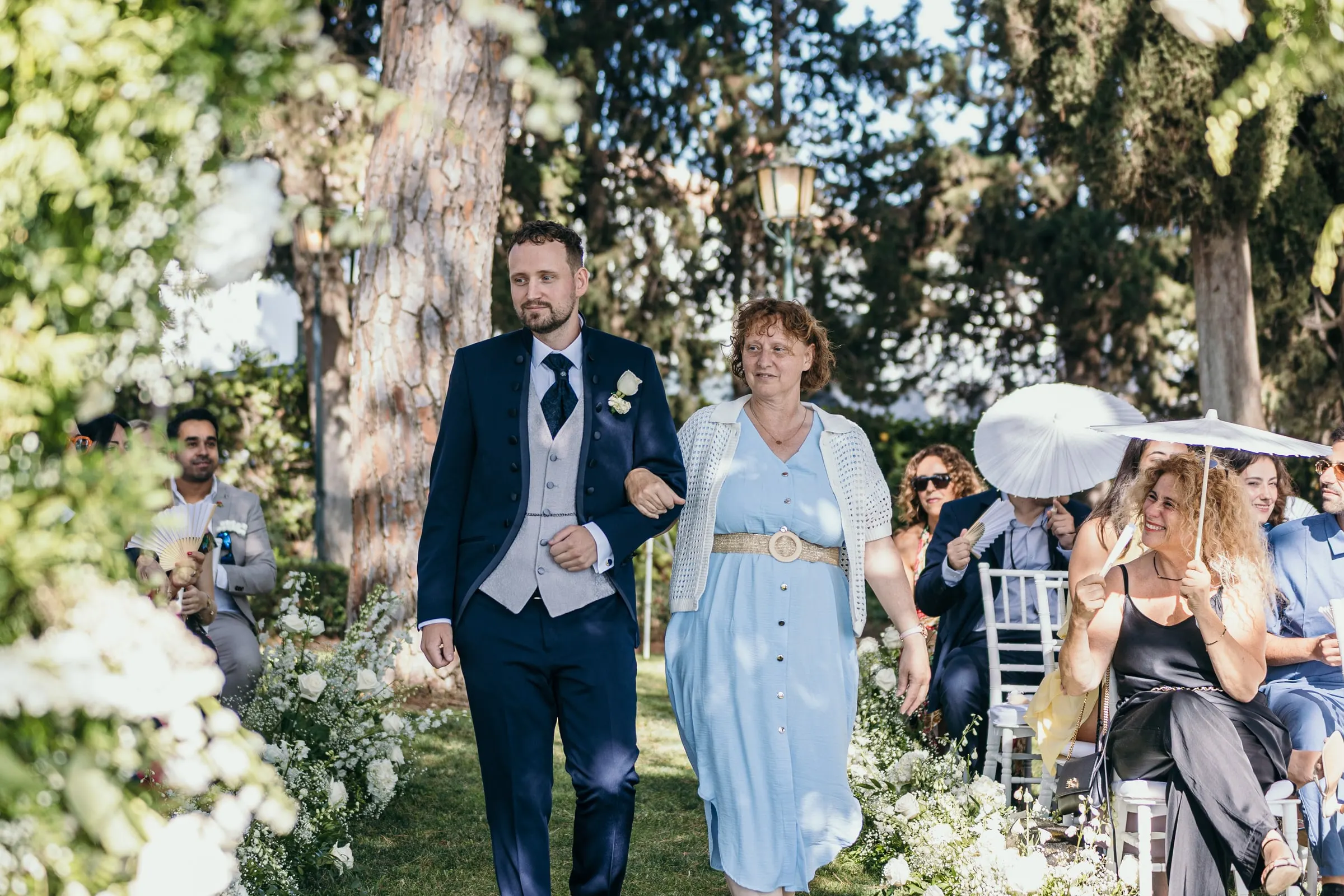 Vincent recorre el pasillo del jardín con una mujer de vestido azul claro mientras los invitados miran desde sus sillas