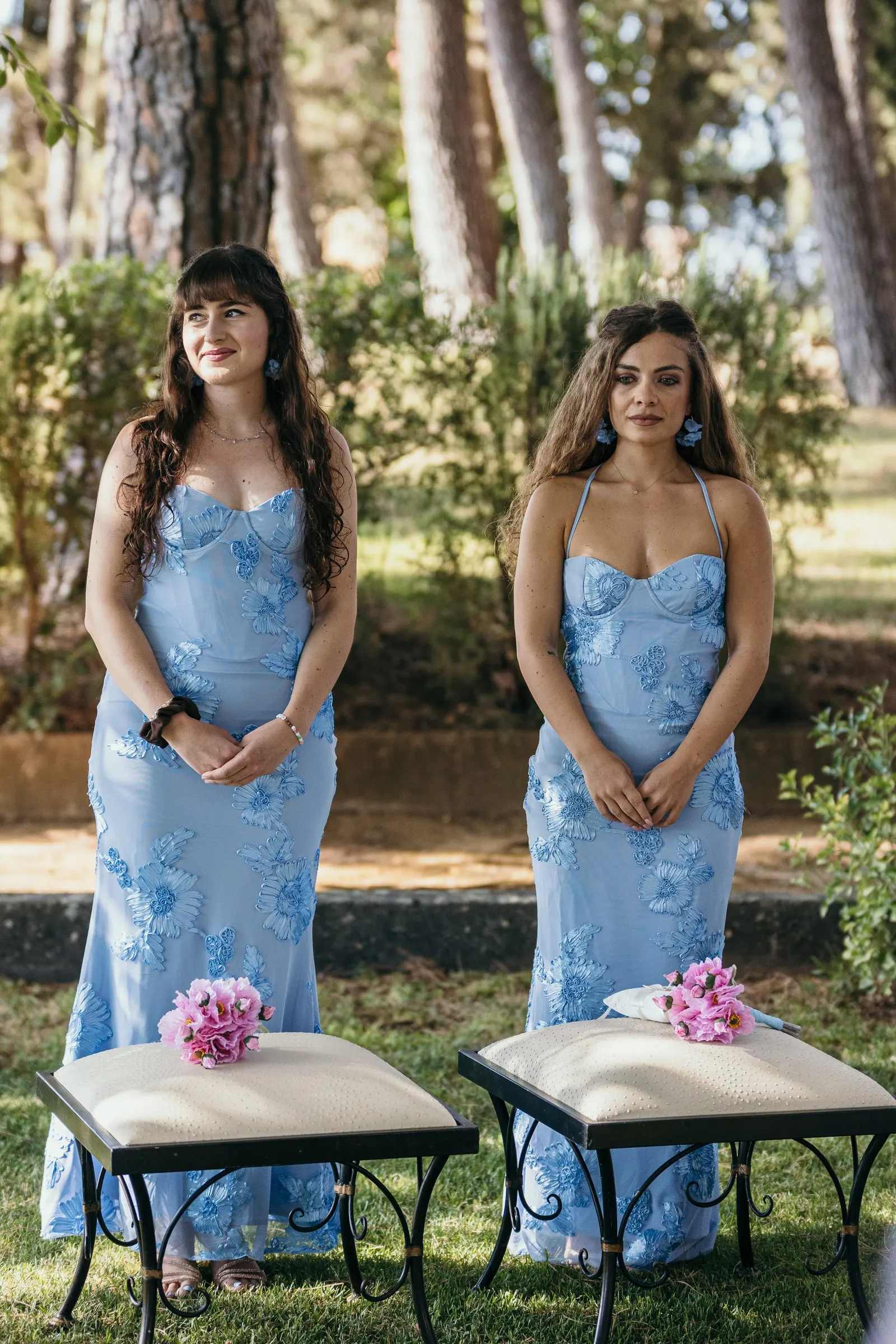 Dos damas de honor con vestidos azules de flores a juego, de pie junto a dos taburetes blancos en el jardín