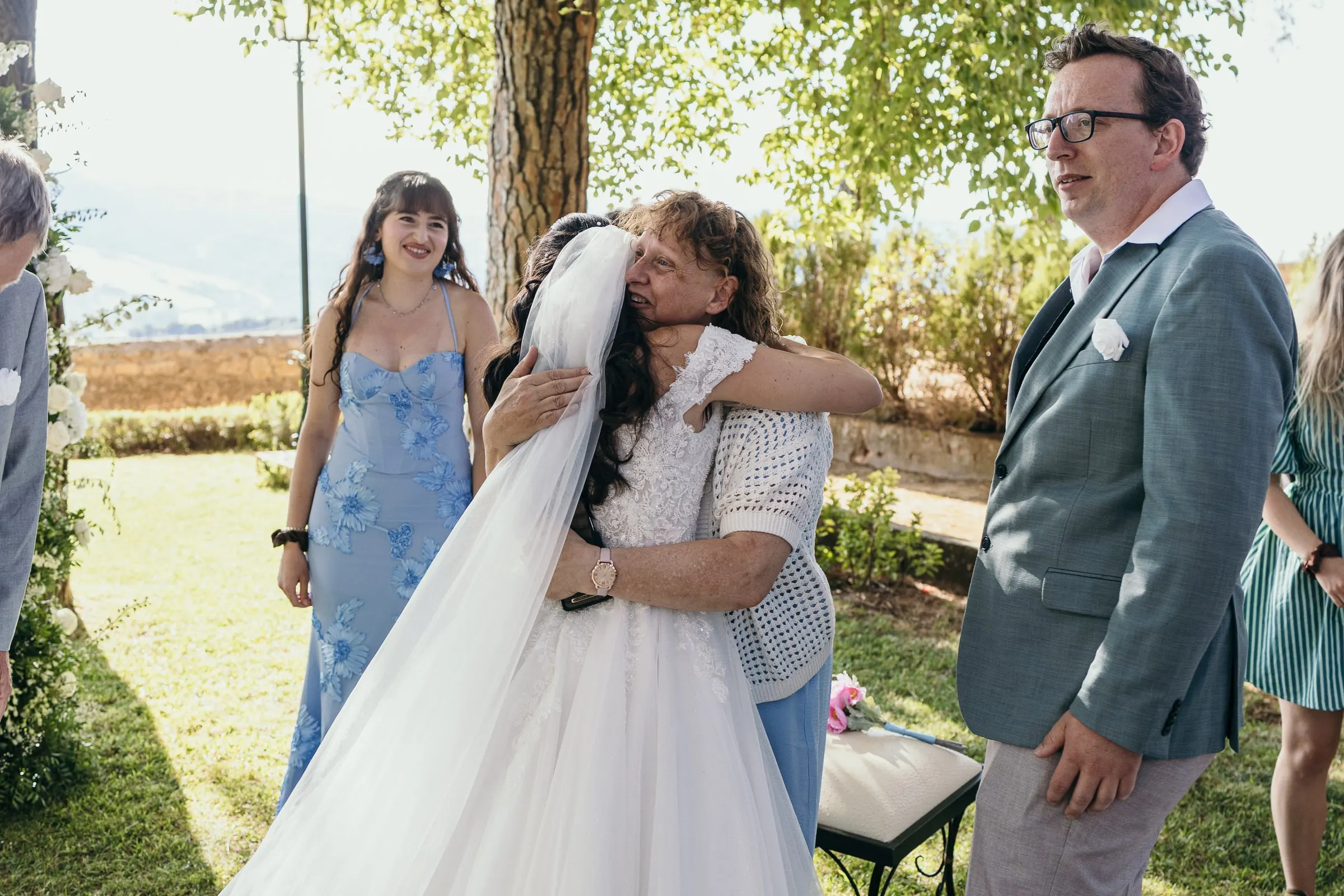 Una invitada abraza a Sara en el césped, con una dama de honor de azul al lado