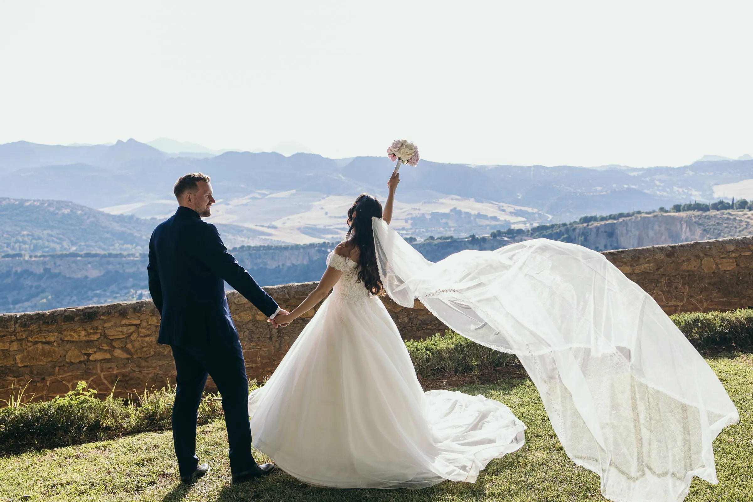 Sara raises her bouquet as her veil flies in the wind above the valley, Ronda