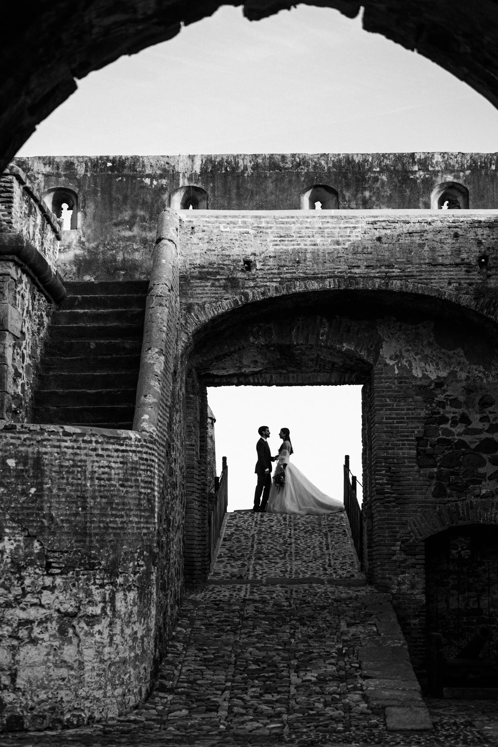 Black and white of the couple framed by a stone archway in an old fortress, Cortijo San Francisco, Estepona