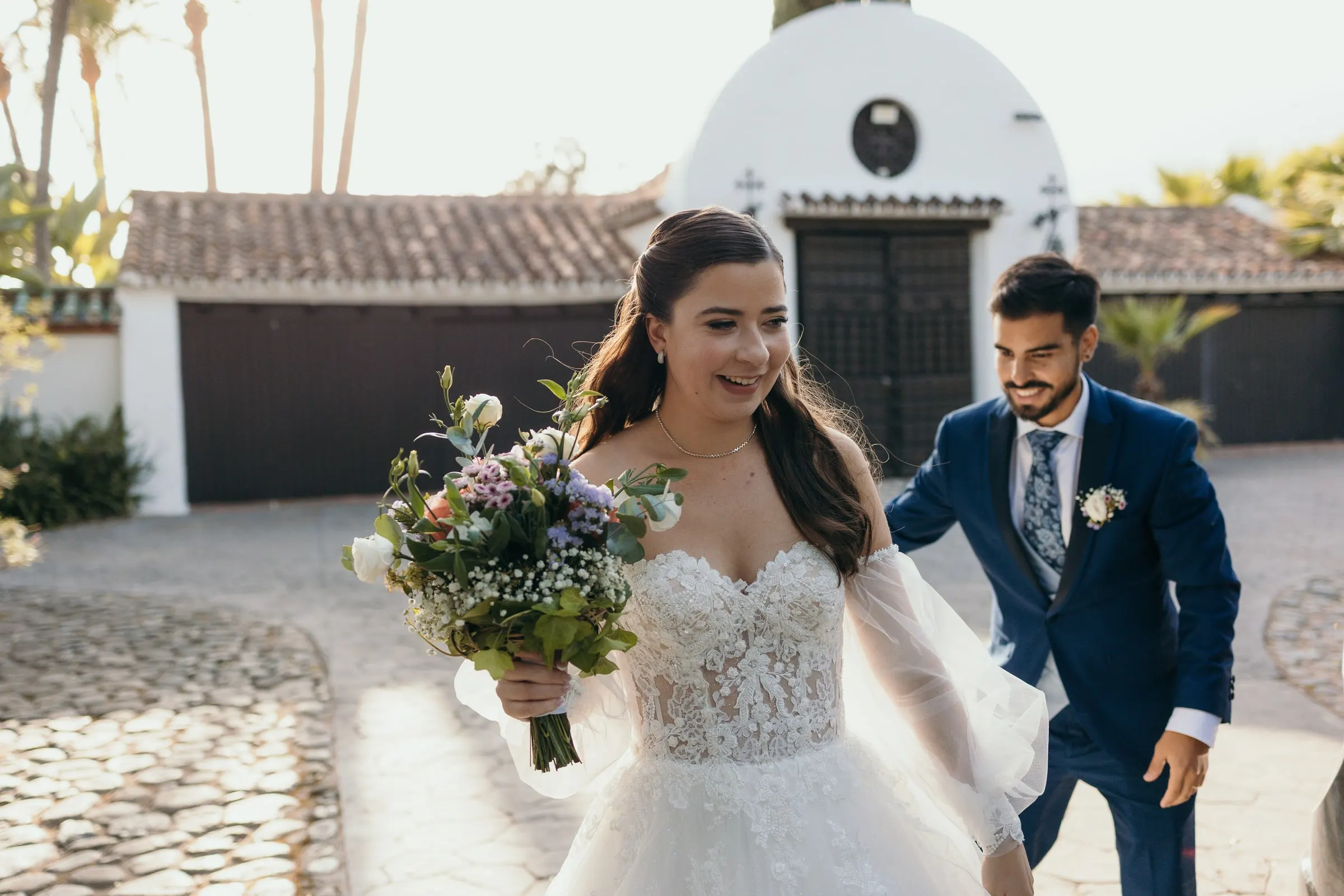 Bride smiling with her bouquet as the groom walks behind her across the courtyard, Cortijo San Francisco, Estepona