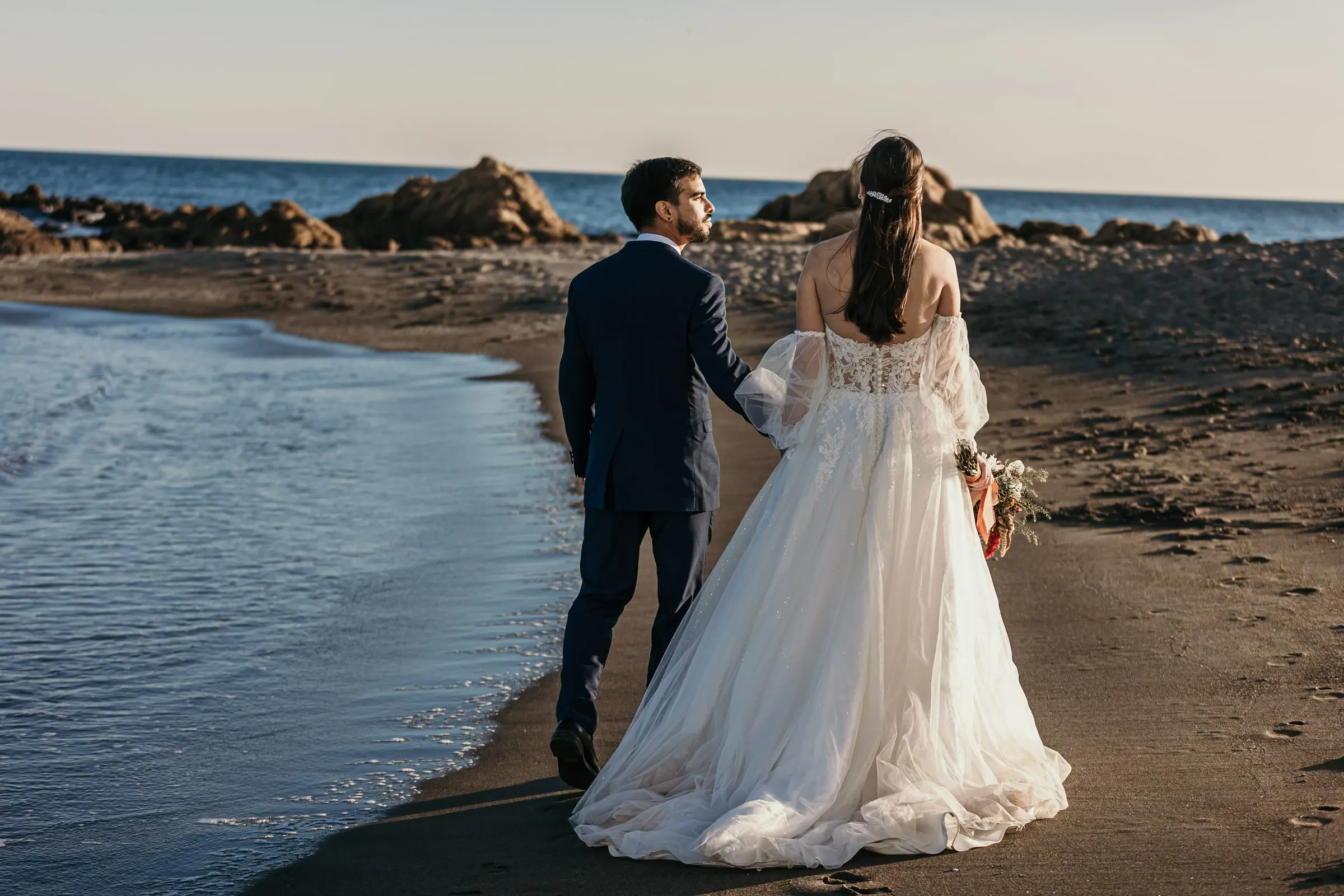 Los novios caminan de la mano por la orilla, vistos de espaldas, Cortijo San Francisco, Estepona