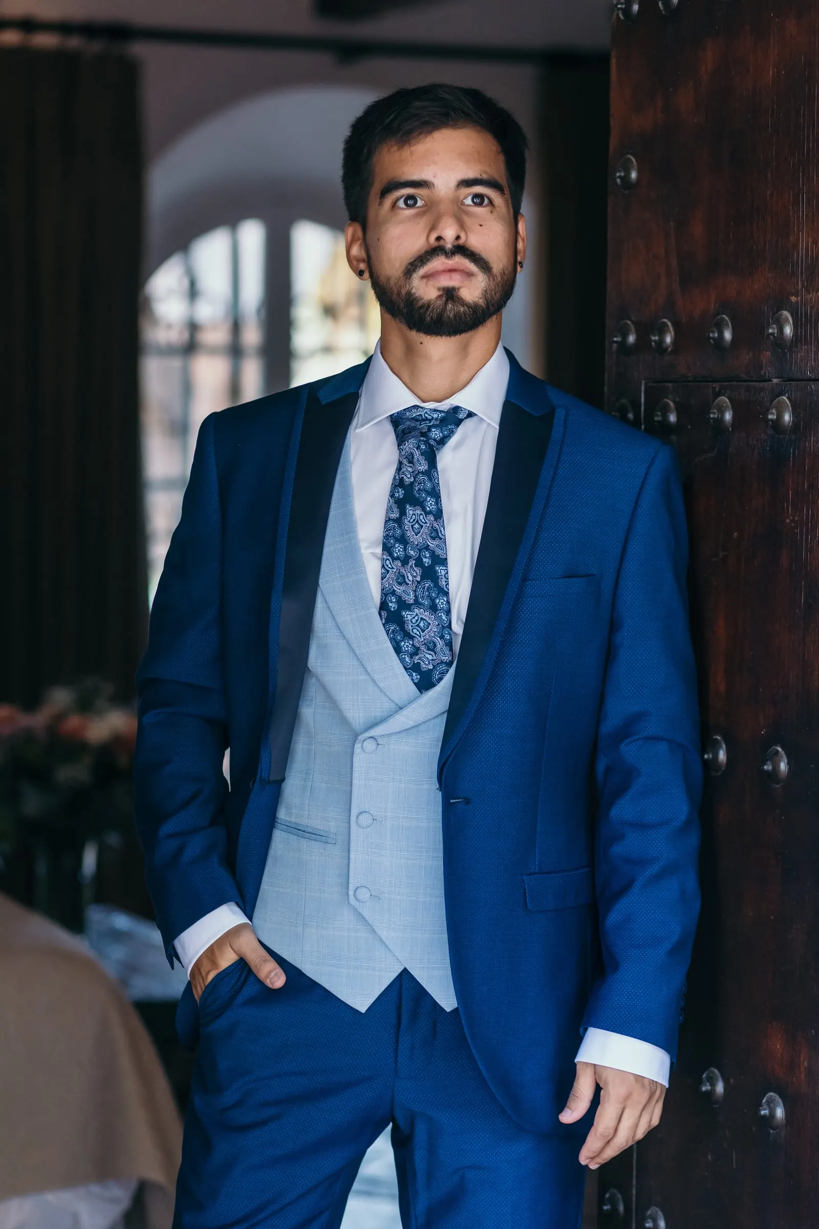 El novio posa con traje azul y chaleco gris junto a un portón de madera con clavos, Cortijo San Francisco, Estepona