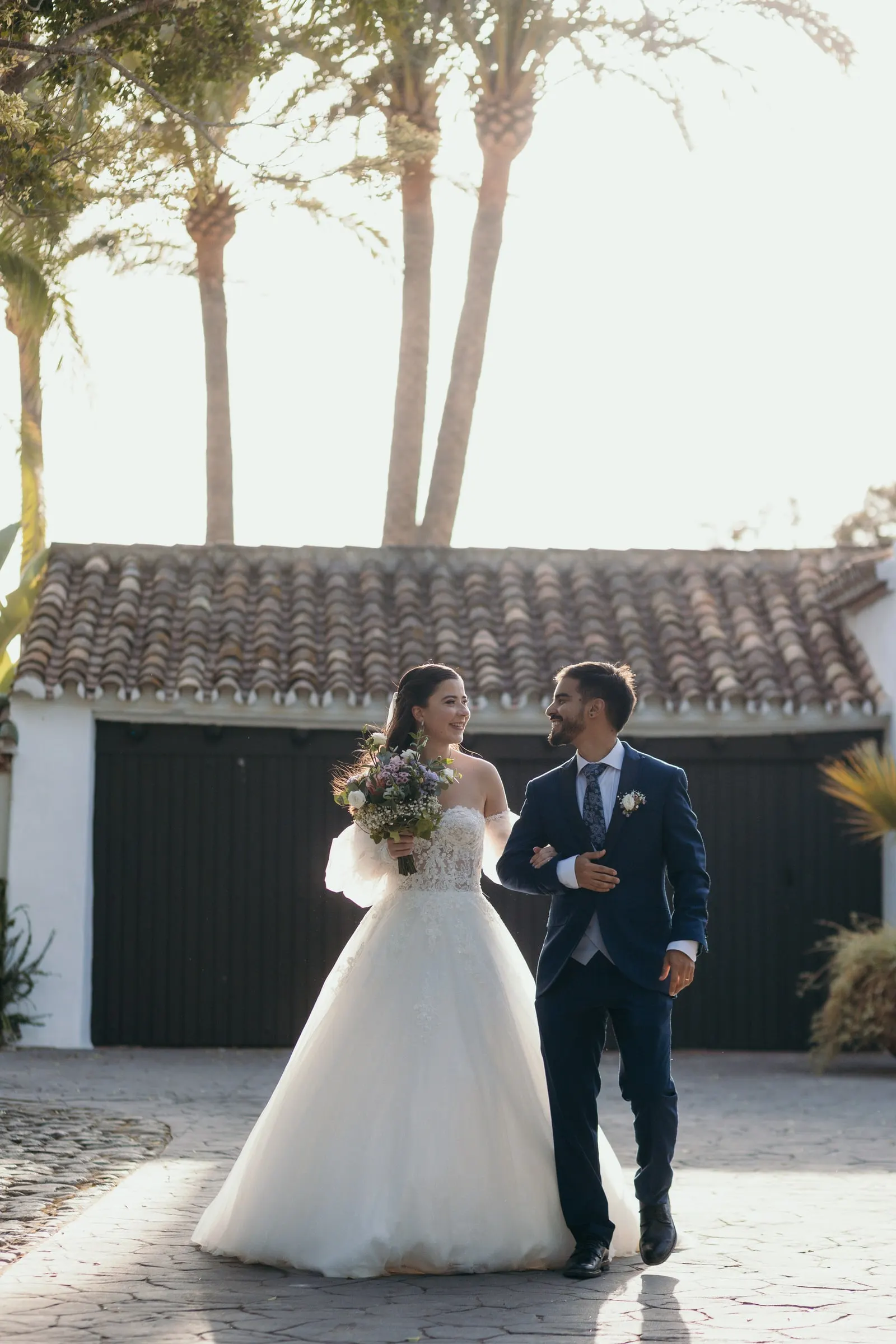 Los novios caminan del brazo por un patio bajo las palmeras, Cortijo San Francisco, Estepona