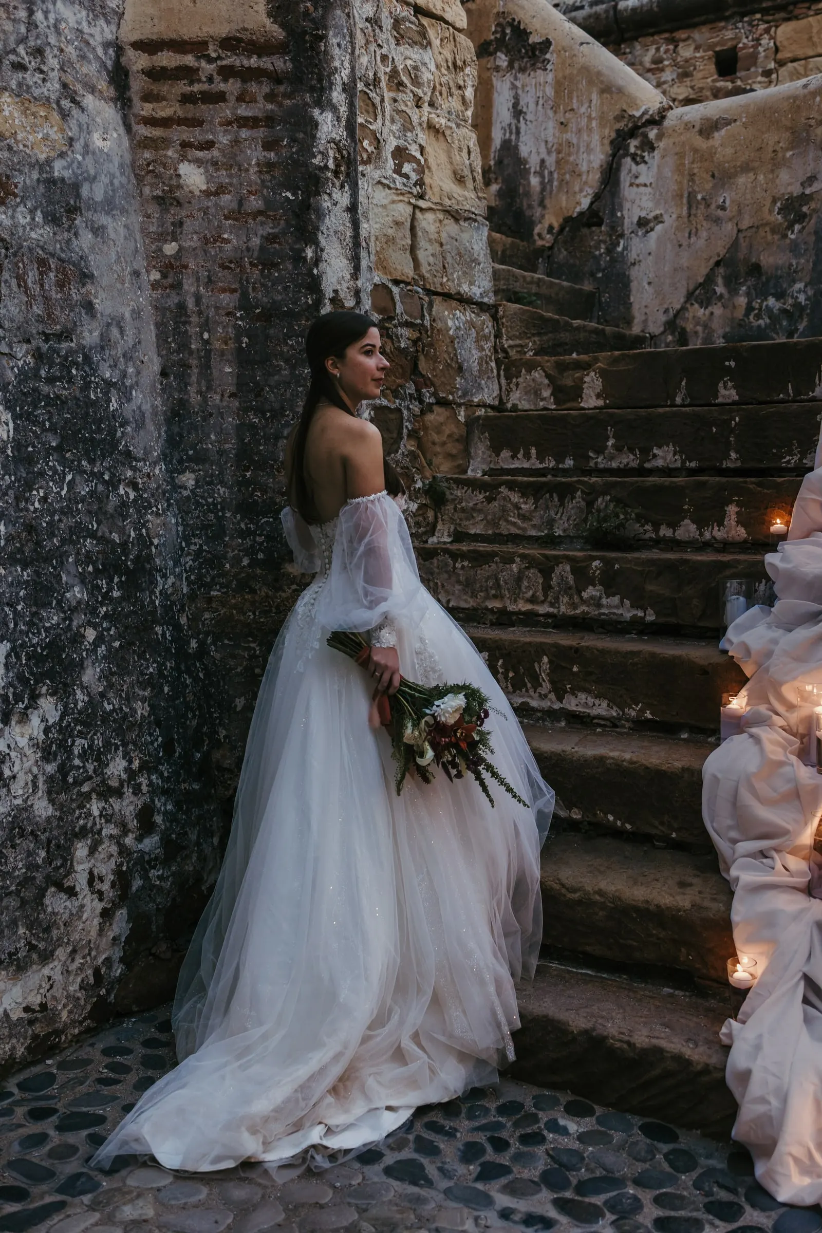 La novia mira por encima del hombro al pie de una antigua escalera de piedra, Cortijo San Francisco, Estepona
