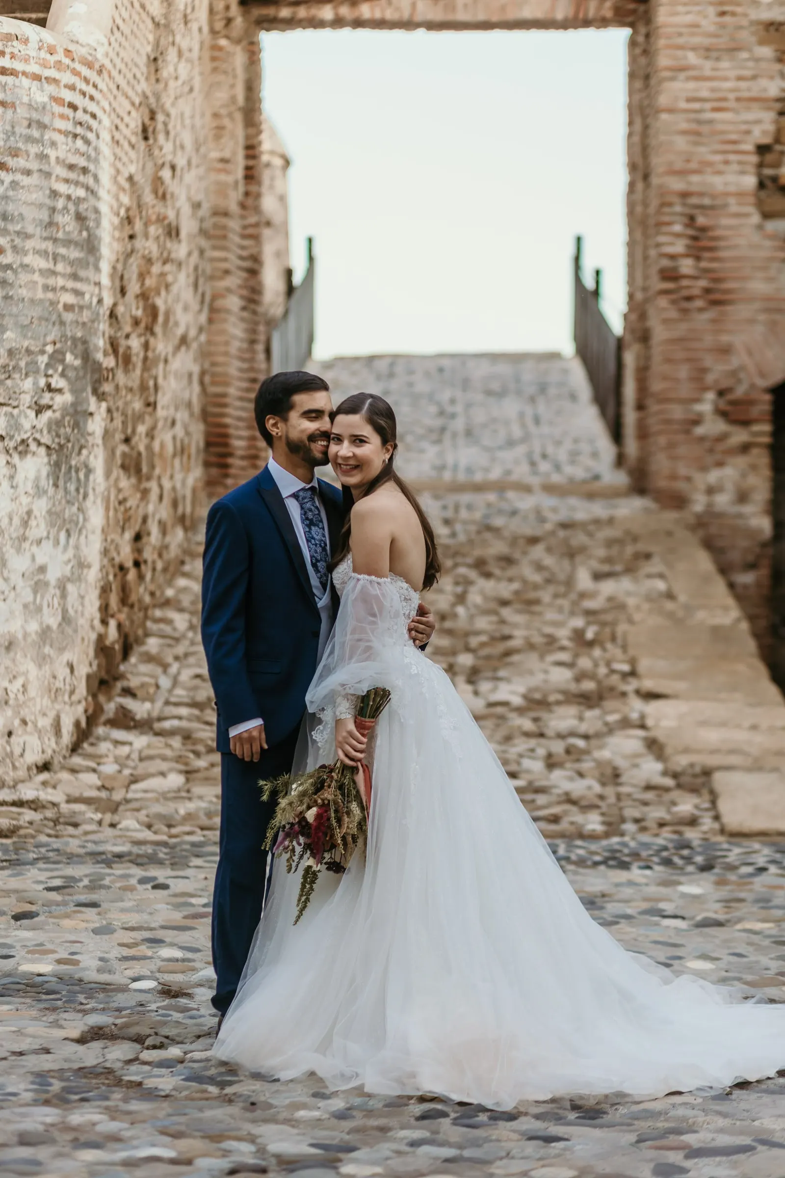 Los novios abrazados y sonrientes en un camino empedrado entre muros antiguos, Cortijo San Francisco, Estepona