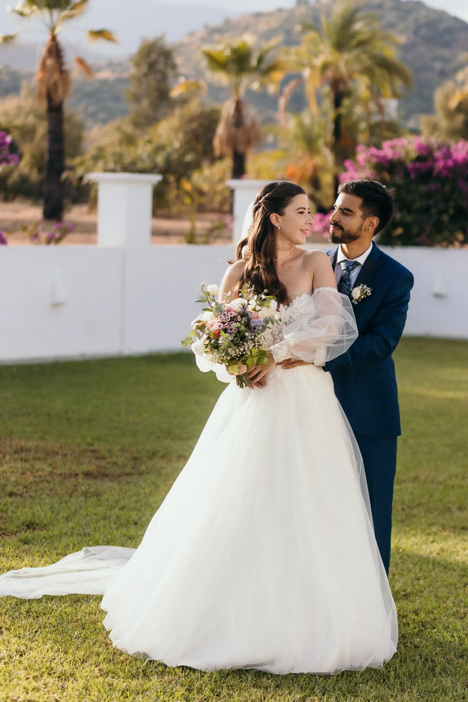 El novio abraza a la novia por detrás en el césped, con las colinas al fondo, Cortijo San Francisco, Estepona