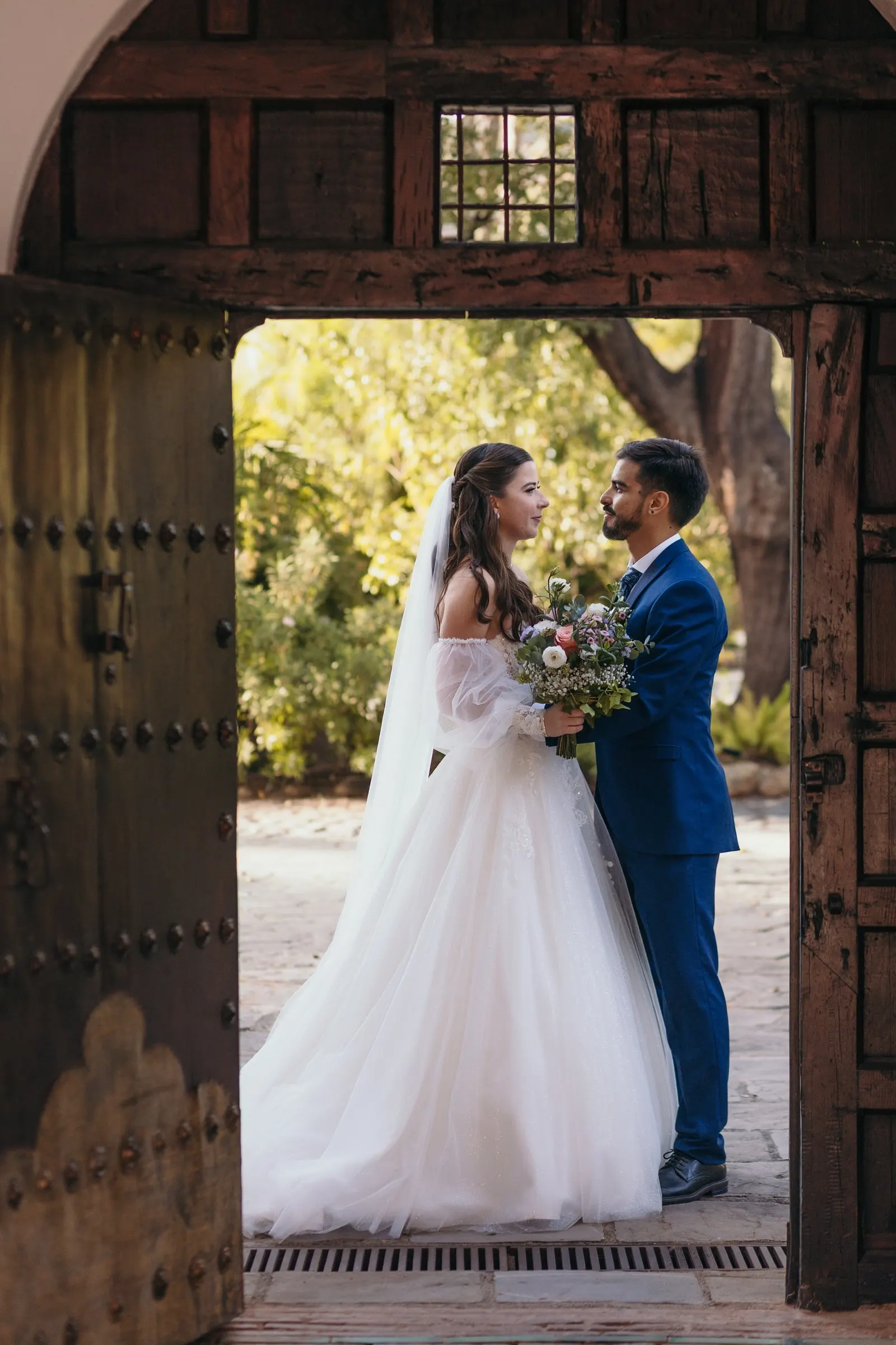 Los novios frente a frente bajo un portón de madera abierto al jardín, Cortijo San Francisco, Estepona