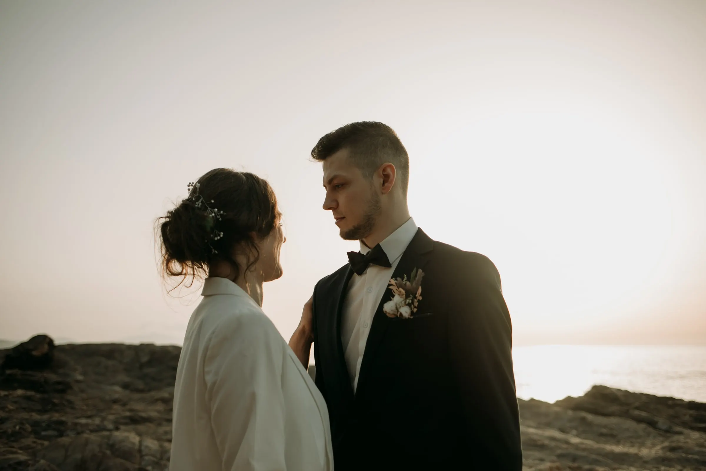La novia con blazer blanco sonríe al novio de esmoquin ante el cielo brumoso del atardecer