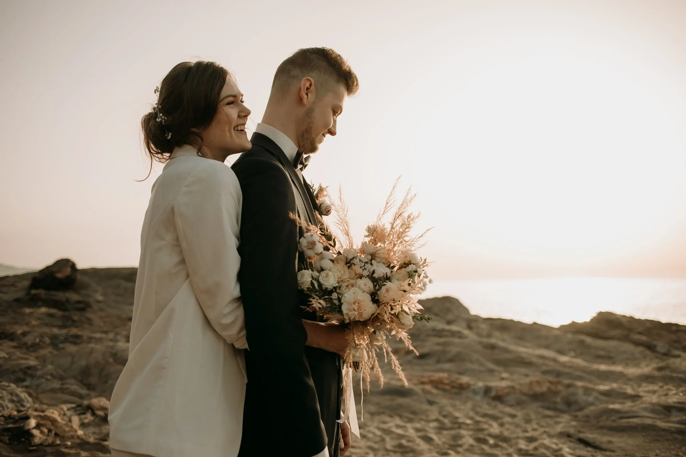 La novia abraza al novio y le sonríe sobre las rocas junto al mar