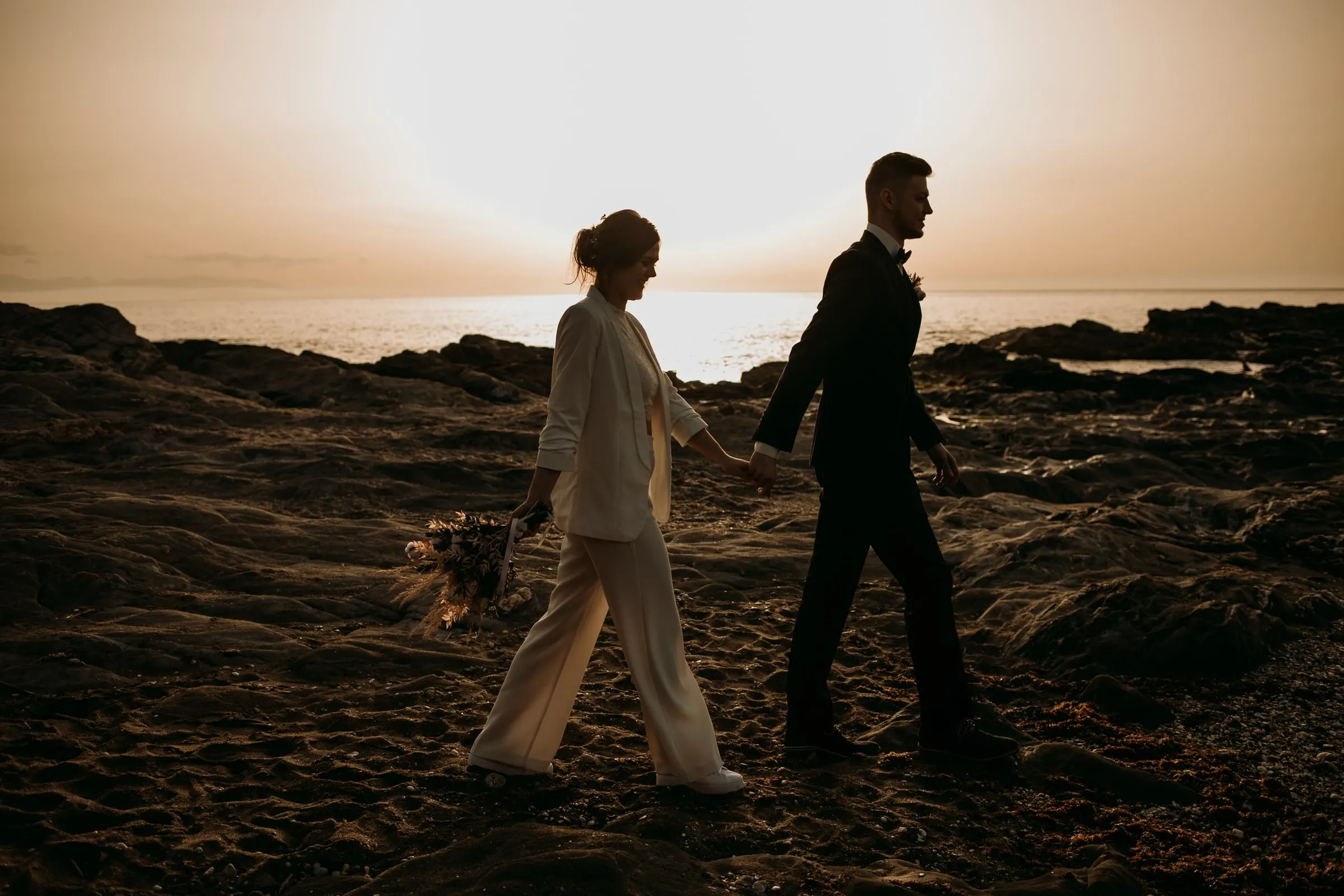 Los novios caminan de la mano sobre rocas oscuras con el mar brillando detrás