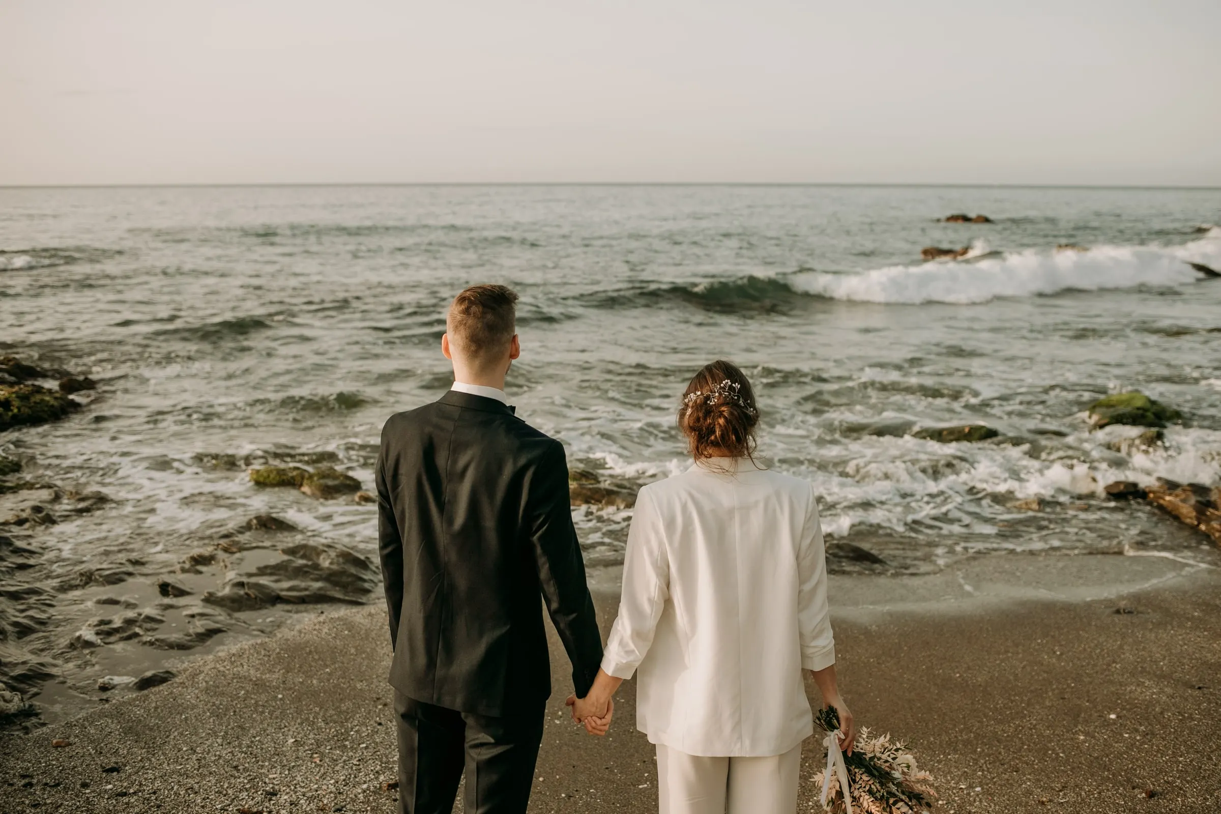 Los novios de espaldas, de la mano, mientras las olas llegan a la playa