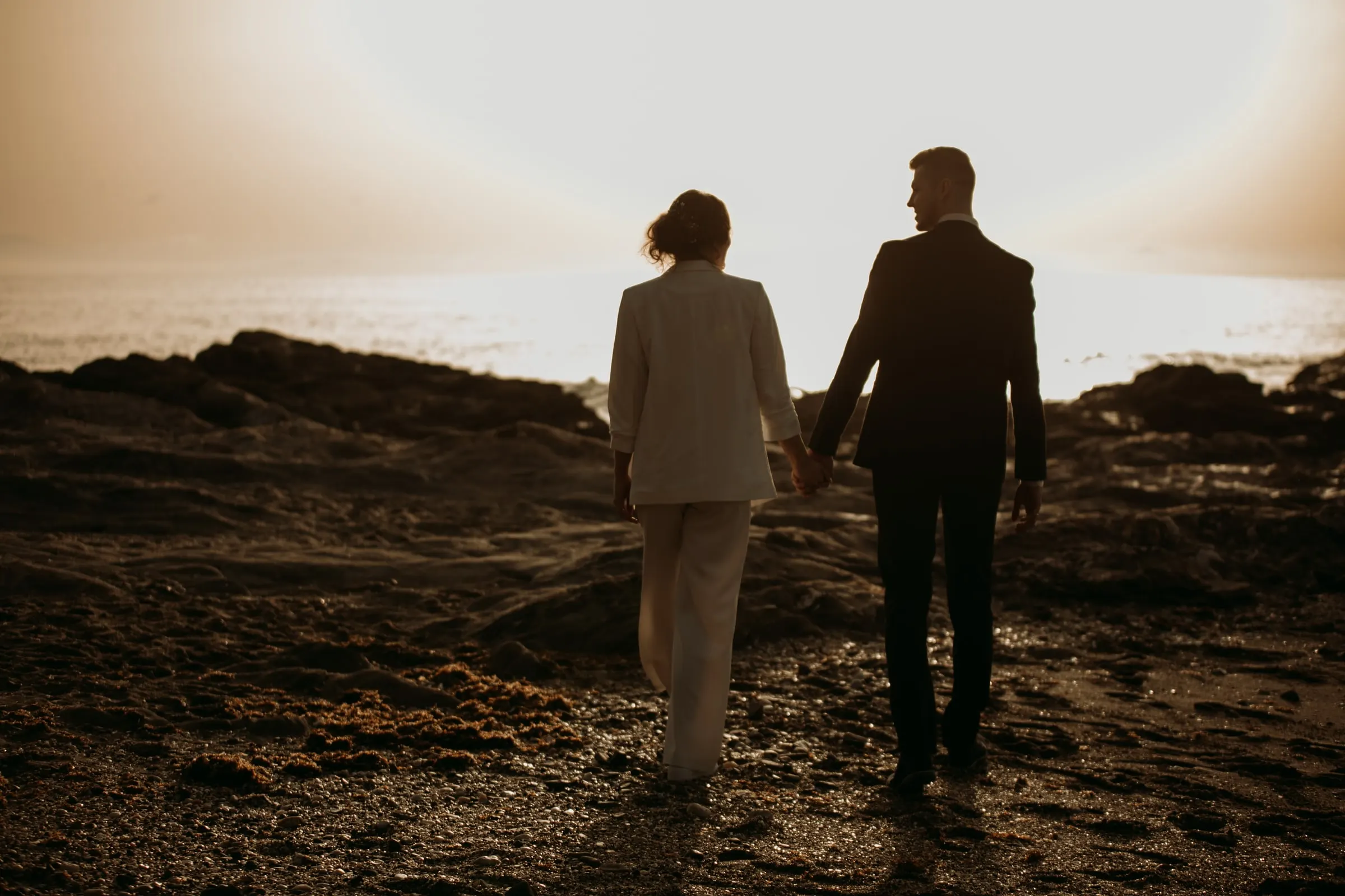 Los novios de espaldas, de la mano, con el sol bajo sobre el mar