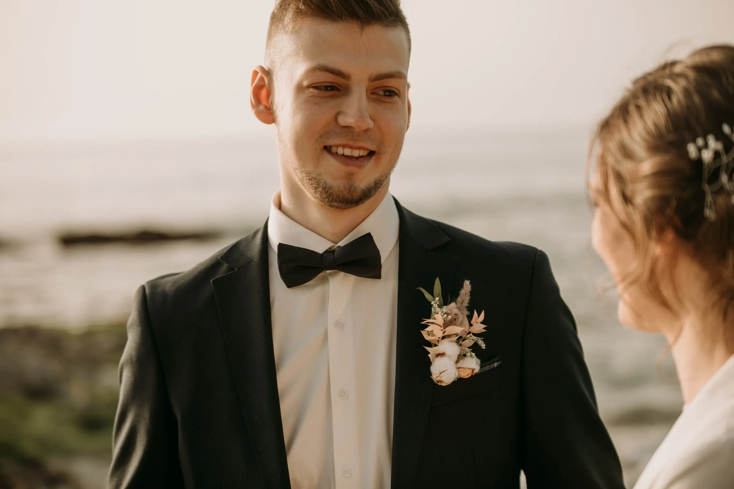 El novio con pajarita sonríe a la novia, con la costa rocosa desenfocada detrás