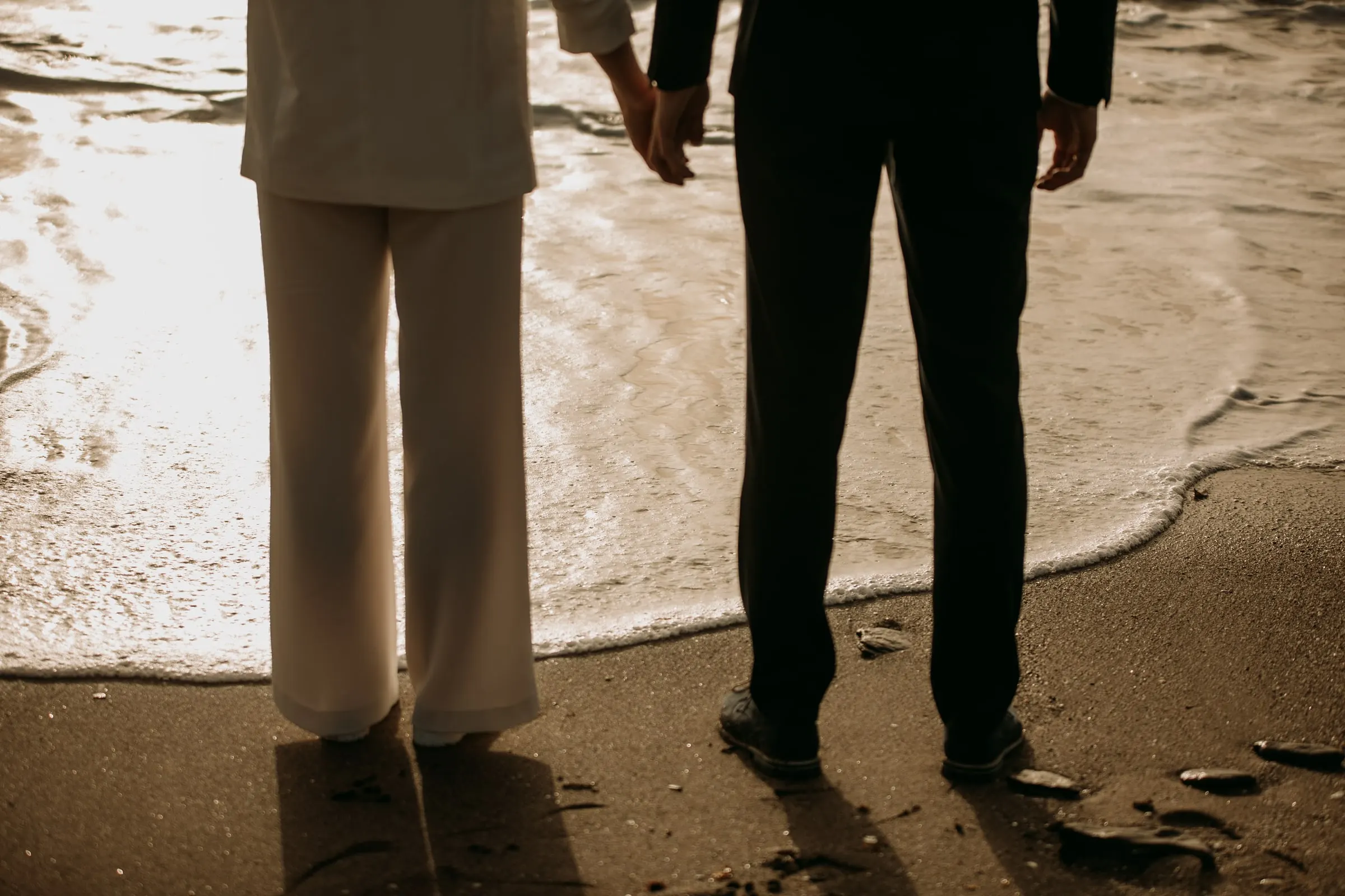 Sus pies al borde de una ola en la arena, pantalón blanco y traje negro
