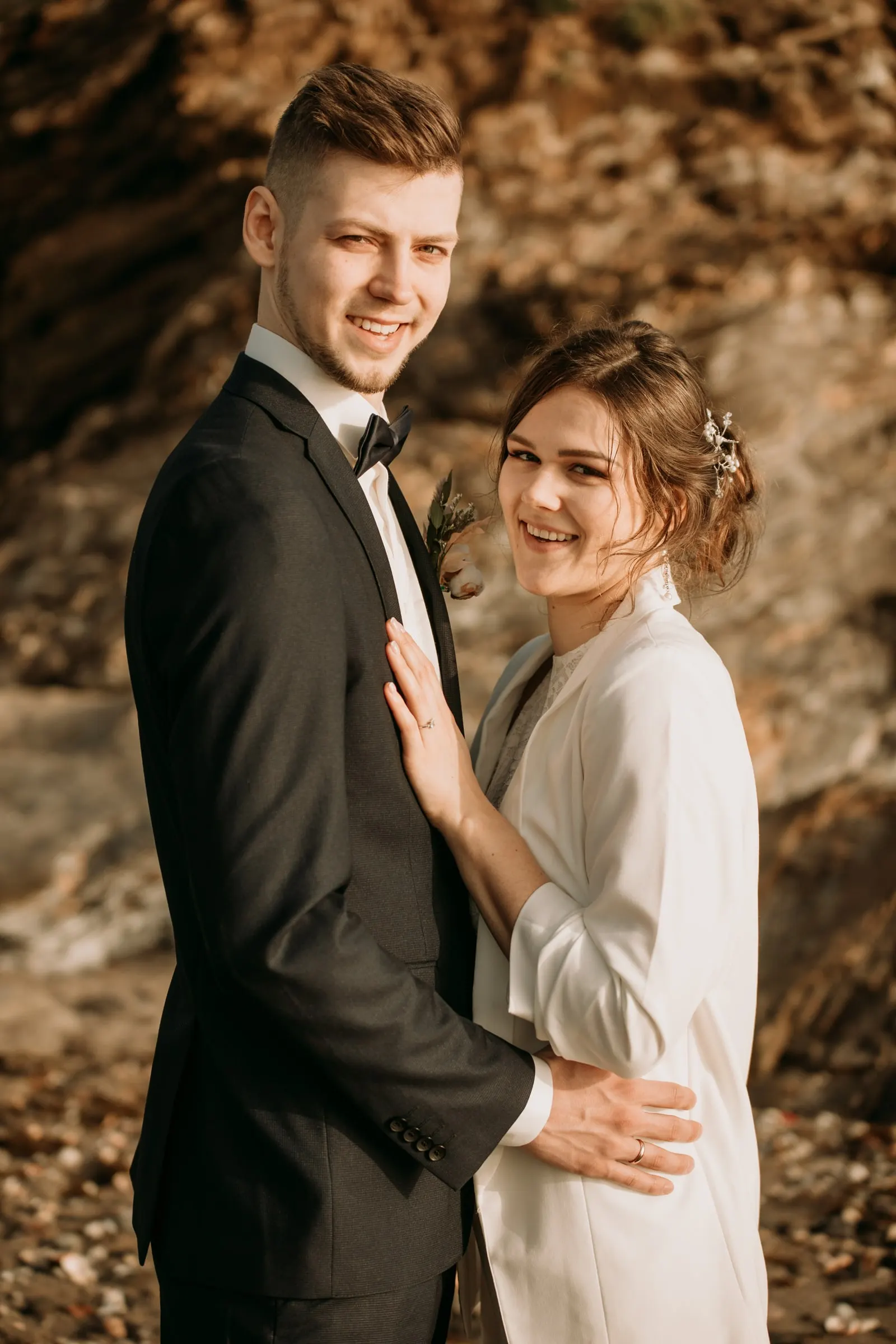 Los novios sonríen a cámara delante de las rocas, con la mano de ella en su pecho