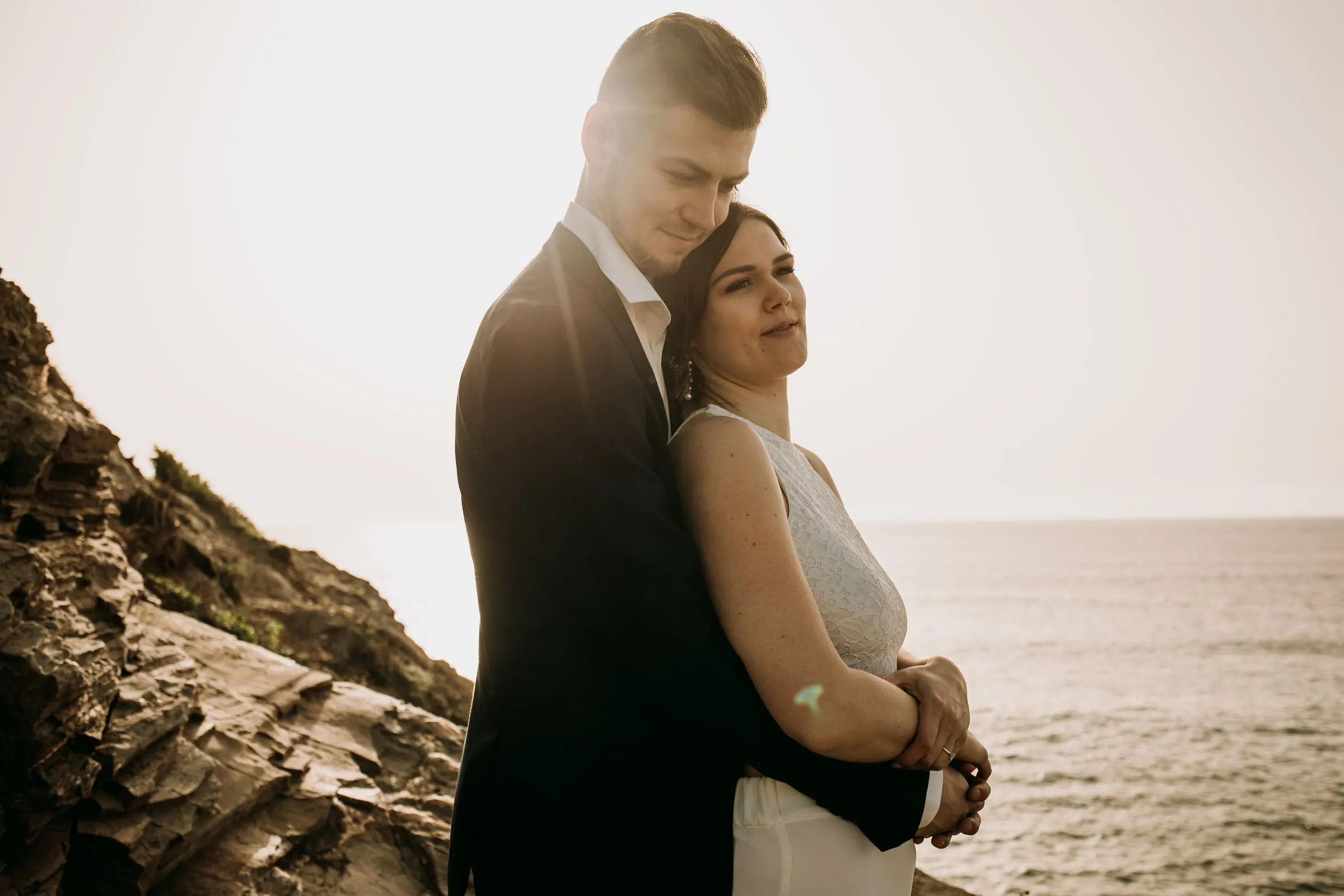 El novio abraza por detrás a la novia al borde del acantilado, con el mar detrás