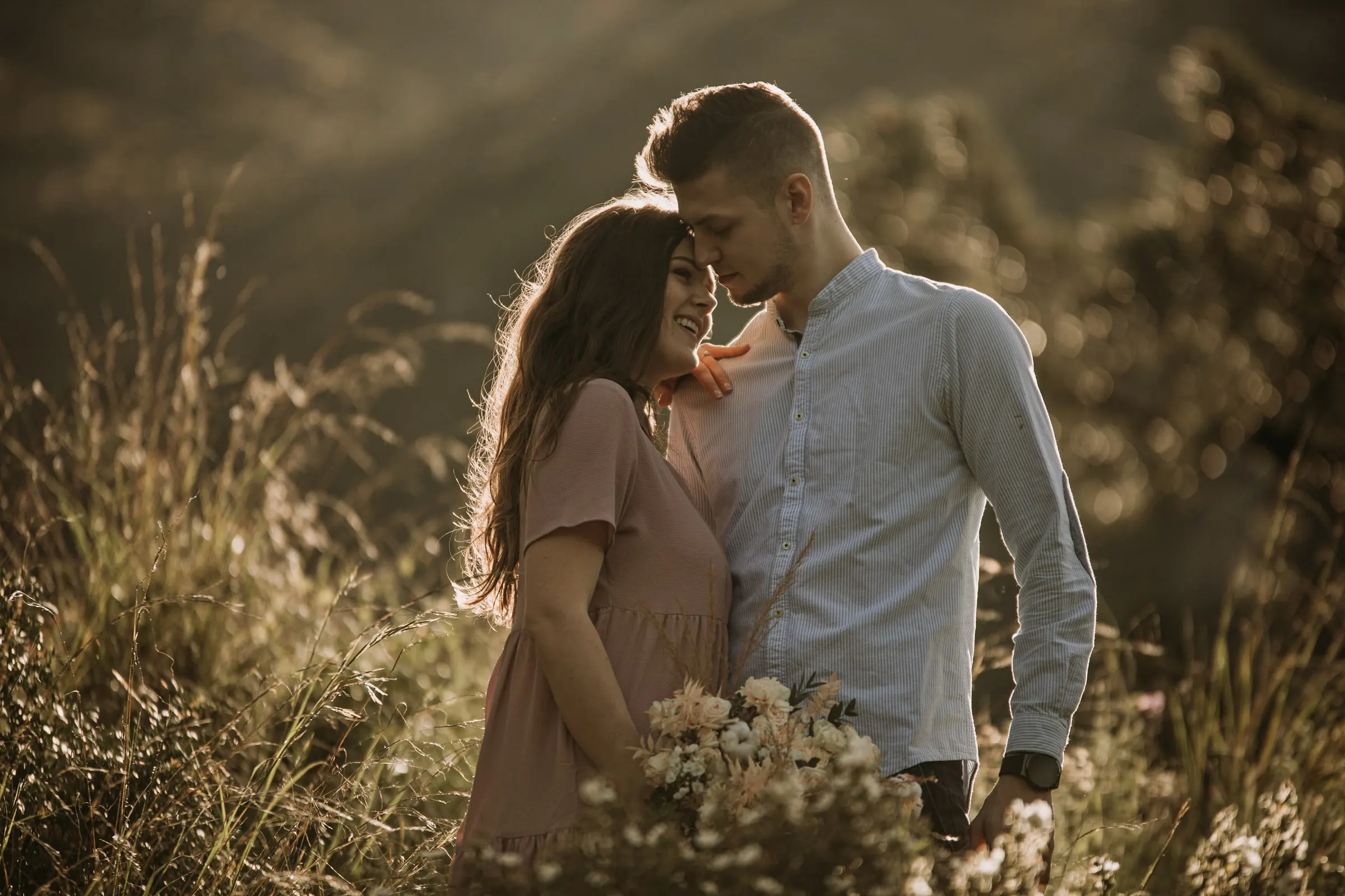 Bride-to-be in a pink dress leaning on her fiancé with her eyes closed in the golden grass