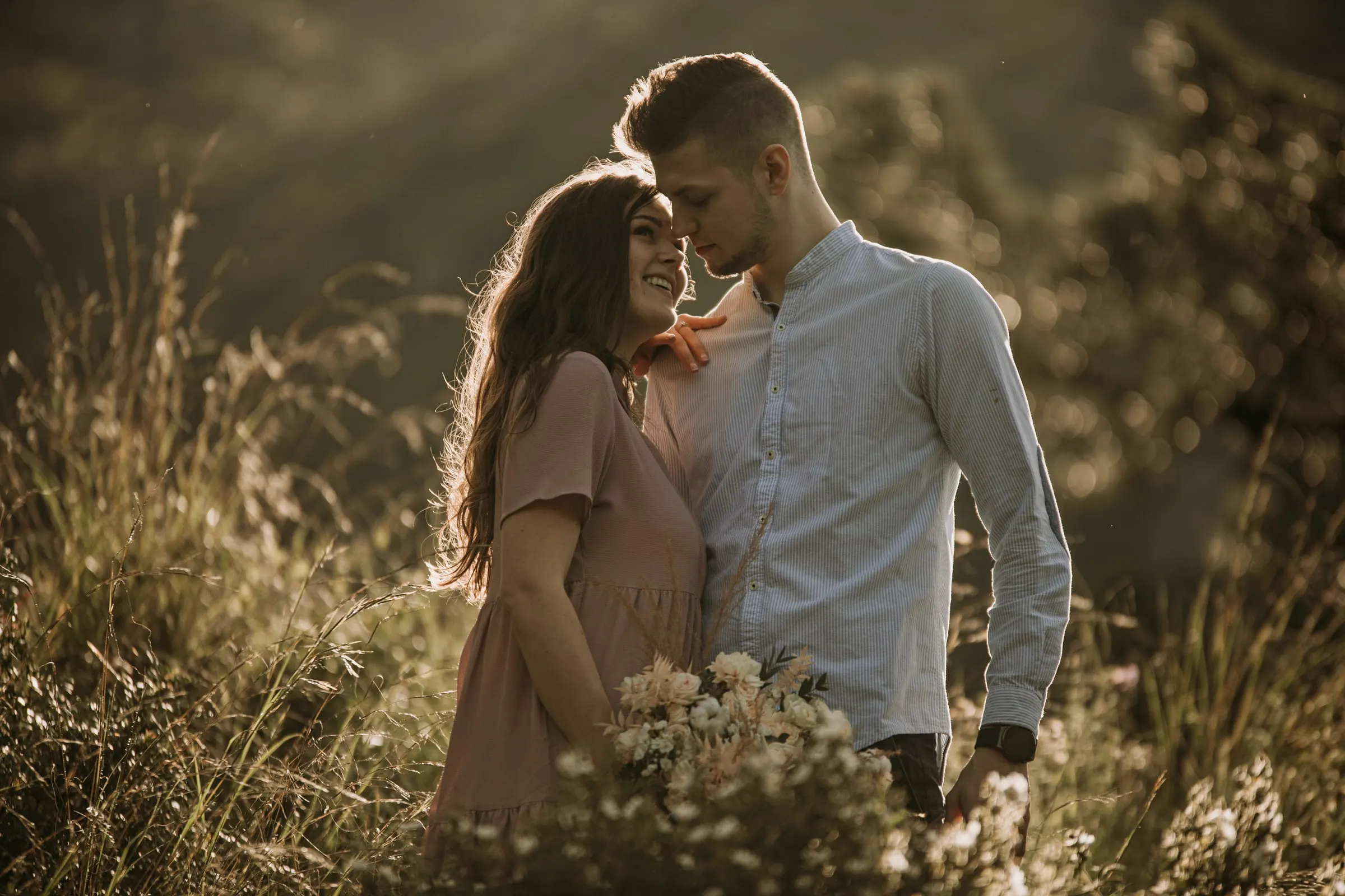 Couple smiling face to face, her hair lit by the sun, a wildflower bouquet in front