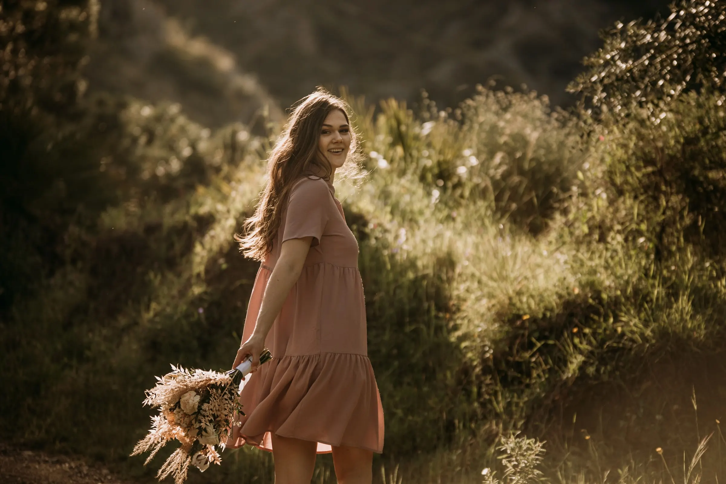Bride-to-be in a pink dress walking through the tall grass and looking back