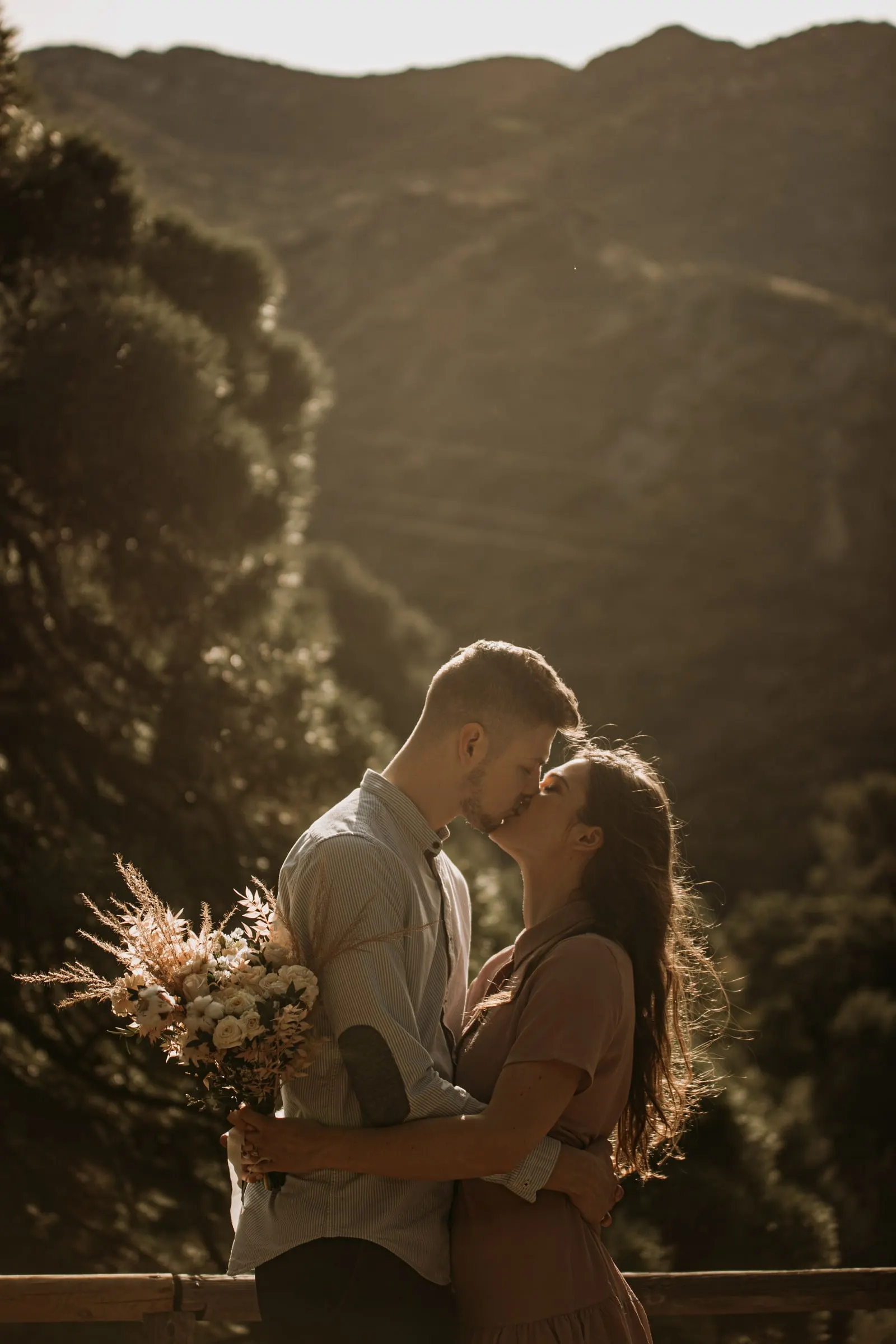 Couple kissing by a wooden railing, pine trees and mountains in soft light