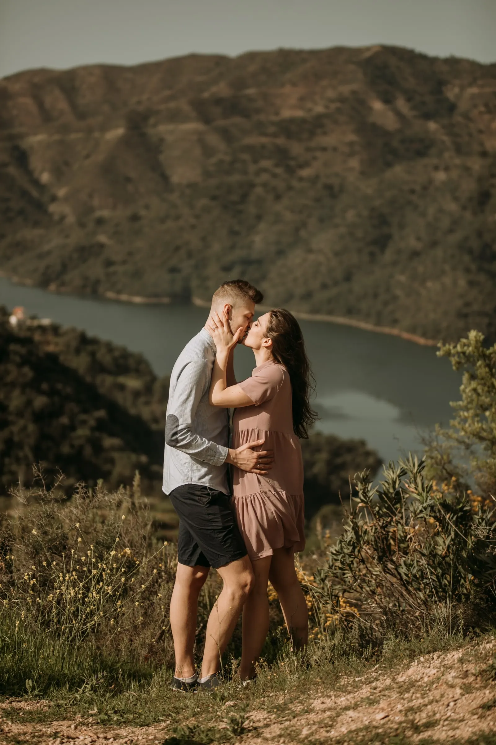 Couple kissing on a hilltop above a reservoir between green mountains, pre-wedding session on the Costa del Sol