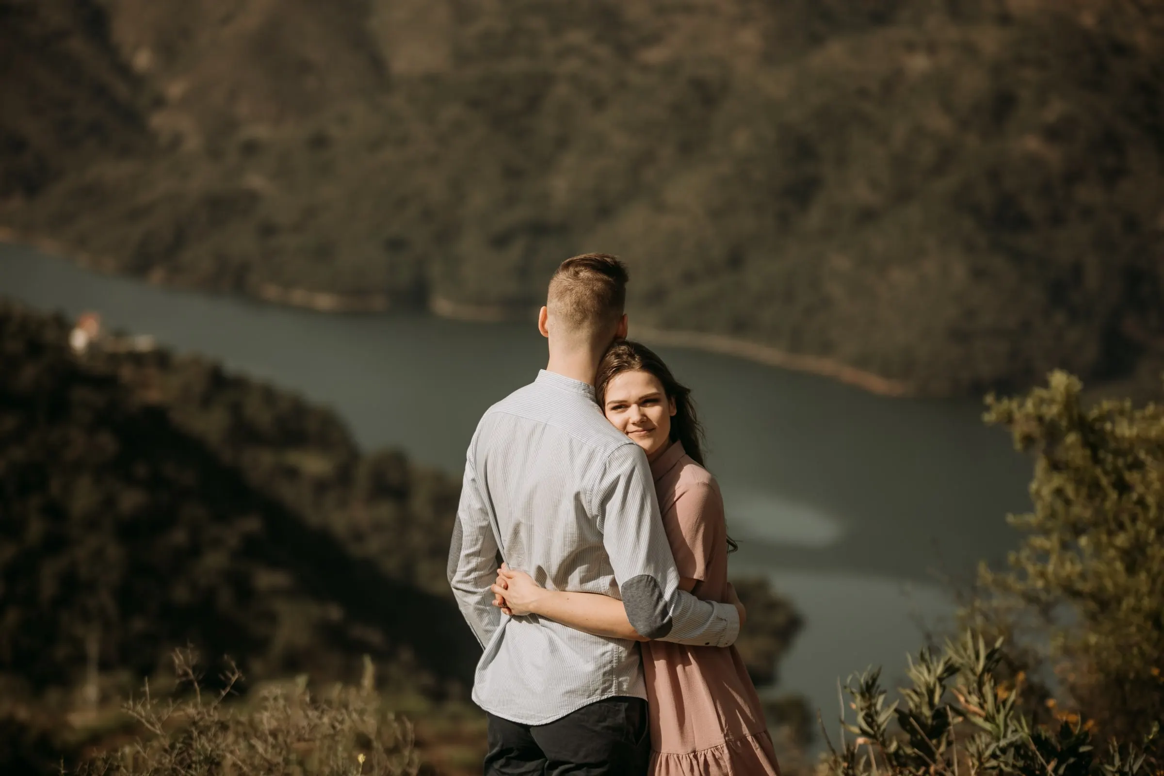 Bride-to-be smiling over her fiancé’s shoulder, the reservoir blurred below