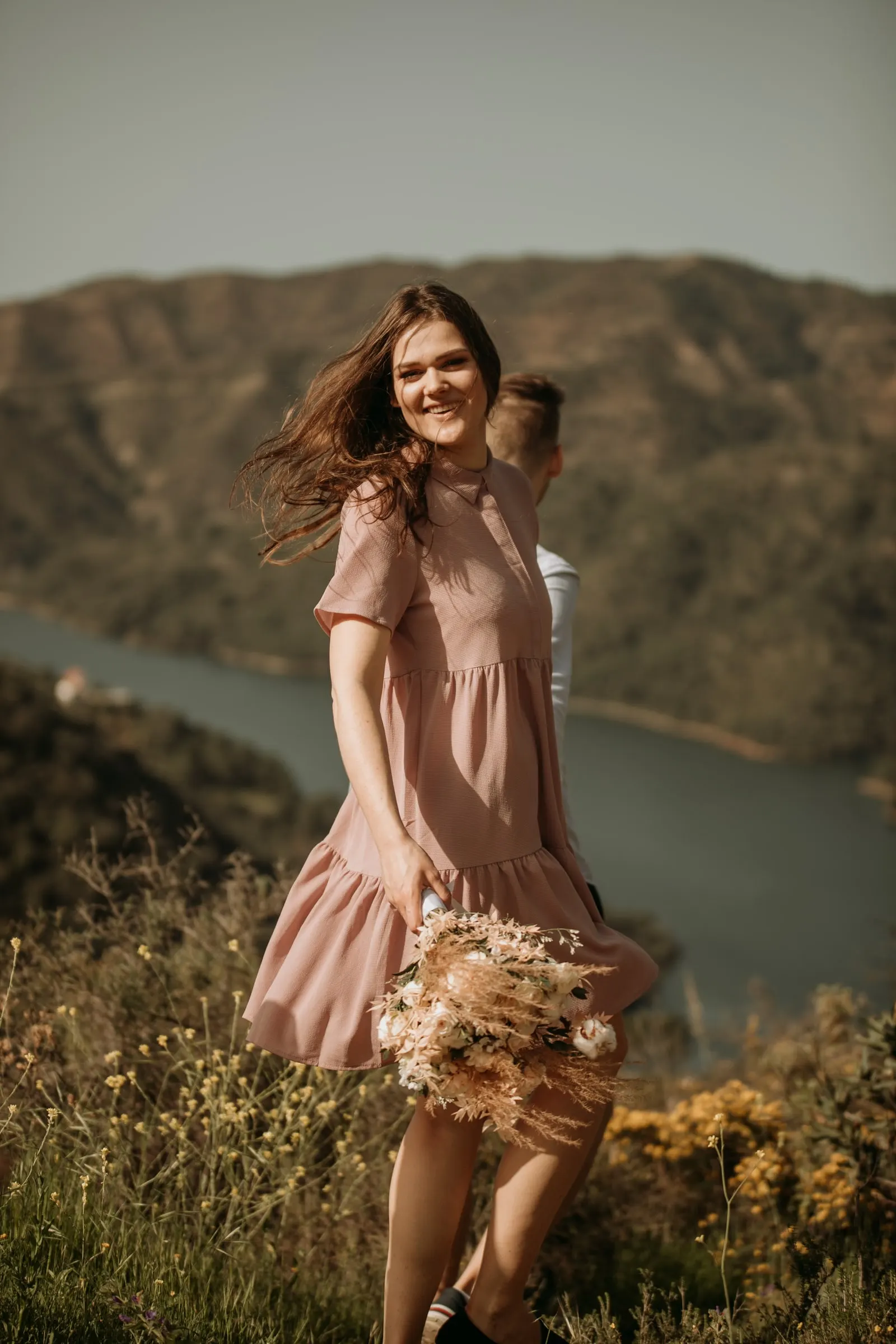 Bride-to-be twirling in her pink dress, hair in the wind, the reservoir behind